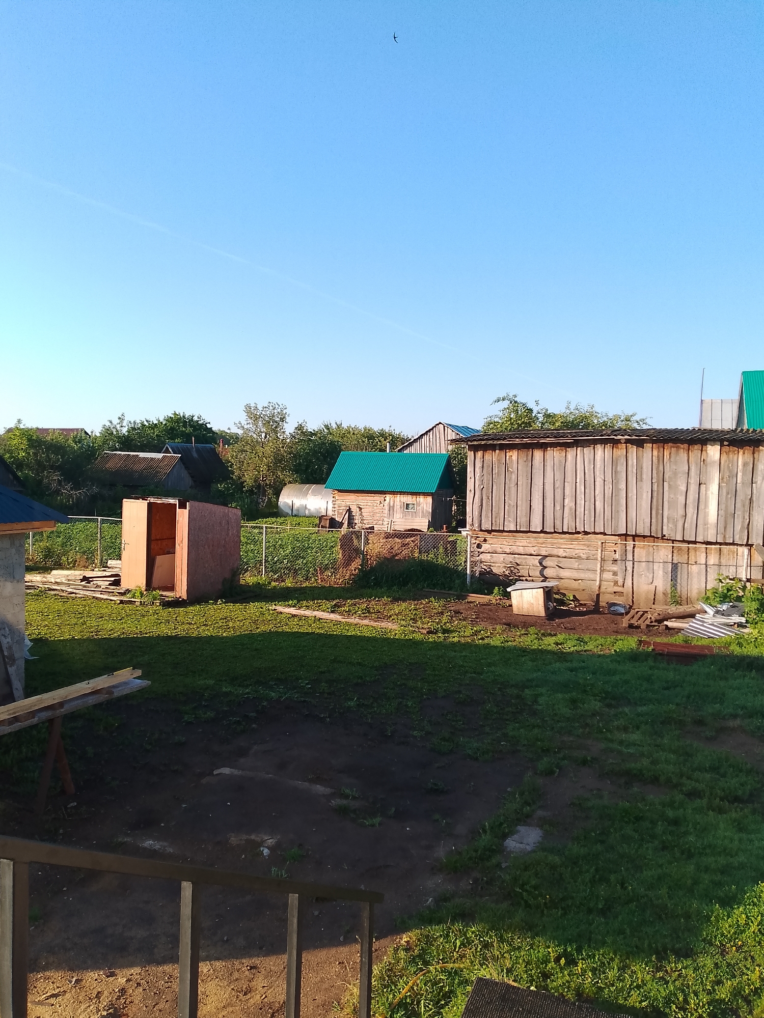 The image shows a backyard scene in Kranokholmskiy, Russia, on a clear day with a bright blue sky. In the foreground, there is a dark wooden railing and some wooden planks resting on a support. The middle ground features a grassy yard with some muddy patches, a small wooden shed under construction, and a larger wooden structure with a corrugated metal roof. Behind these structures, a chain-link fence separates the yard from a garden area with rows of plants and a greenhouse. Several other buildings with various roof types, including green and grey corrugated metal, are visible in the background, surrounded by trees and vegetation. A single bird is captured in flight in the upper left portion of the sky. The scene appears to be a residential or dacha property, suggesting a rural or semi-rural setting.