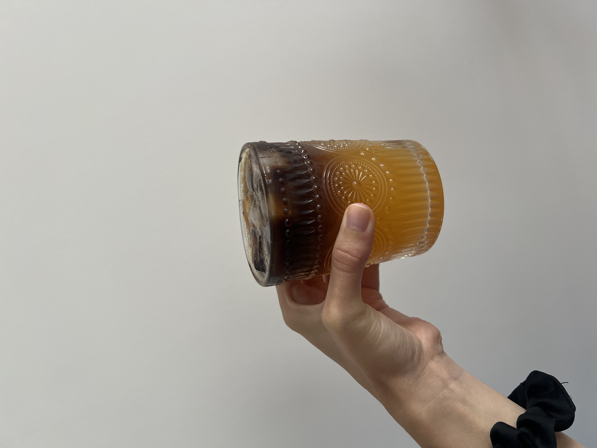 A hand holds a glass tumbler at an angle against a plain white background. The tumbler contains a two-layered beverage with dark brown liquid at one end and a lighter orange liquid filling the majority of the glass. Some ice cubes are visible inside the tumbler. The tumbler is made of glass, has a rounded, cylinder shape, and features an embossed design around its circumference. The hand holding the glass has fair skin. A black scrunchie is visible around the wrist of the hand. The background is a uniform, light grey color. The location is impossible to determine based on the image content.