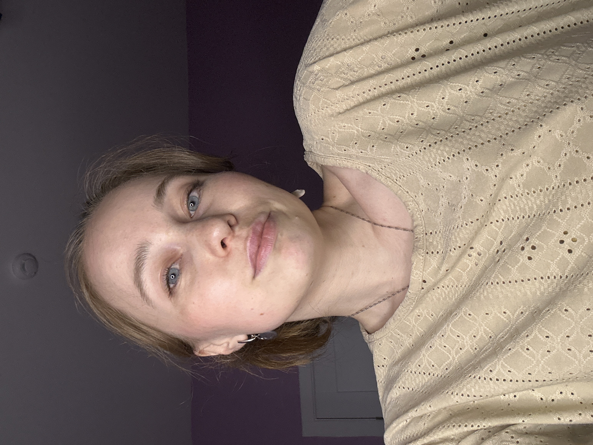 A woman with light skin, light brown hair, and blue eyes is seen in a close-up, selfie-style portrait. She is wearing a light tan, textured top and a delicate silver necklace. Her face shows no apparent makeup. The background consists of a dark gray wall in the upper left and a deep purple wall behind her head and shoulders. A white window frame is partially visible at the bottom edge of the background.
There is no information to suggest the location is Saint Petersburg, Russia, or any city.