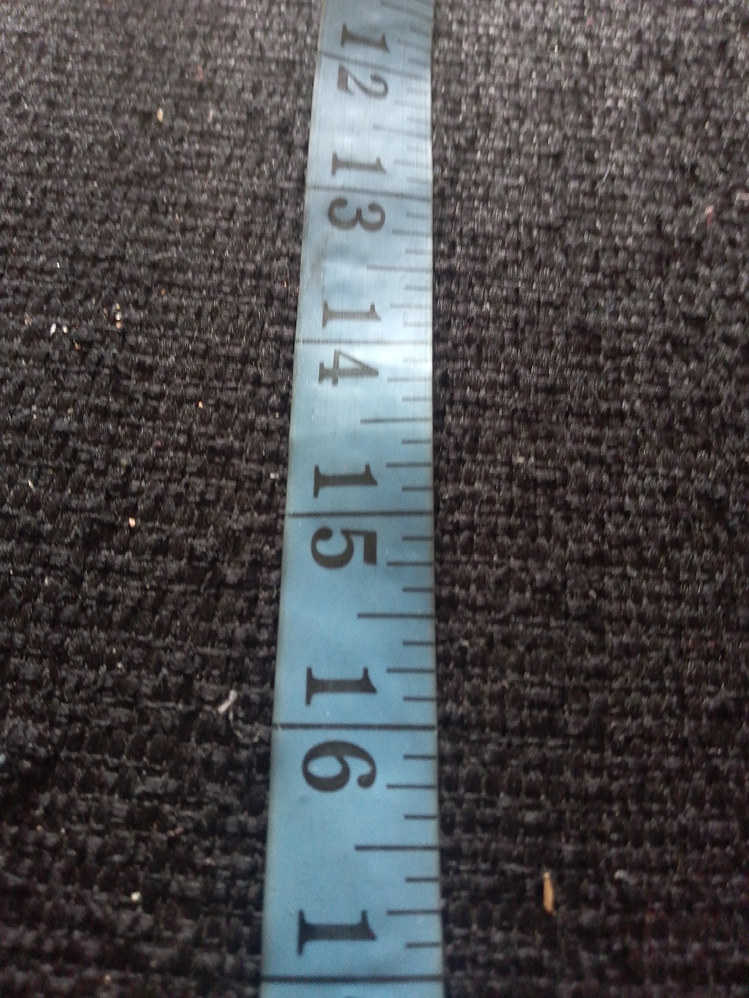 A close-up, slightly angled, static shot shows a blue measuring tape against a dark, textured surface, possibly carpet or fabric. The tape has black markings, indicating measurements. The visible numbers on the tape are 12, 13, 14, 15, 16, and 1. The surface beneath the tape is dark and speckled with very small light-colored particles. The image is entirely focused on the measuring tape and its immediate surroundings. No specific location in Jauro Yinu, Nigeria, can be determined from this image.