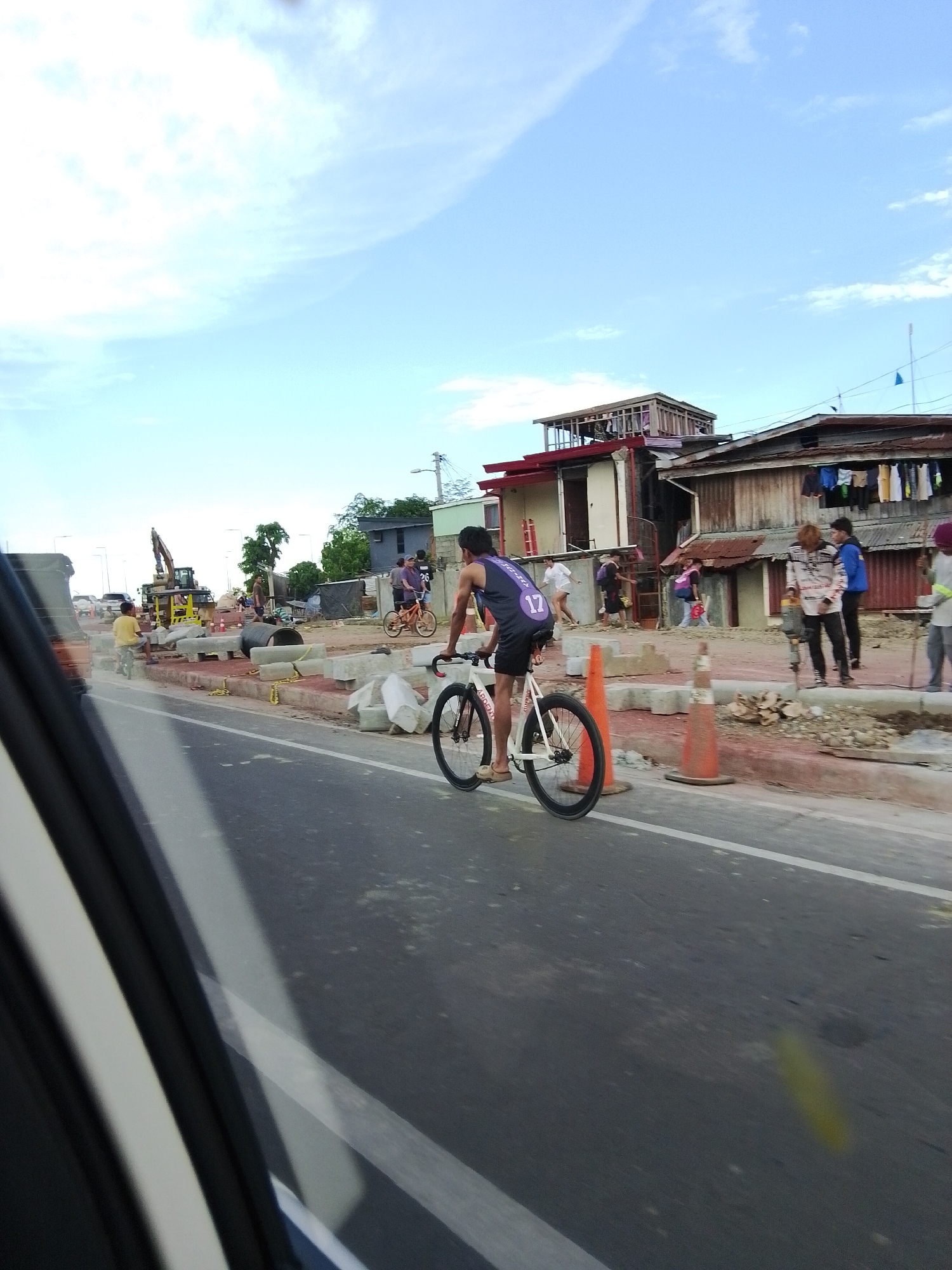 The image shows a street scene in Davao City, Philippines, with ongoing construction and people engaged in various activities. In the foreground, a person is riding a white bicycle with black wheels. The rider is wearing a dark sleeveless shirt with the number "17" on the back and dark shorts. The bicycle appears to be a fixed-gear or single-speed model.
Behind the cyclist, there is a construction site with yellow barriers, concrete blocks, and construction equipment, including a yellow excavator. Several people are present near the construction area, some standing and observing, while others appear to be working. One person is holding a tool, possibly a shovel or a pickaxe.
Further in the background, residential structures with corrugated metal roofs are visible. Laundry is hung out to dry on clotheslines in front of one of the houses. The sky is partly cloudy, with a mix of blue sky and white clouds. The overall impression is a bustling urban environment with elements of daily life and infrastructure development.