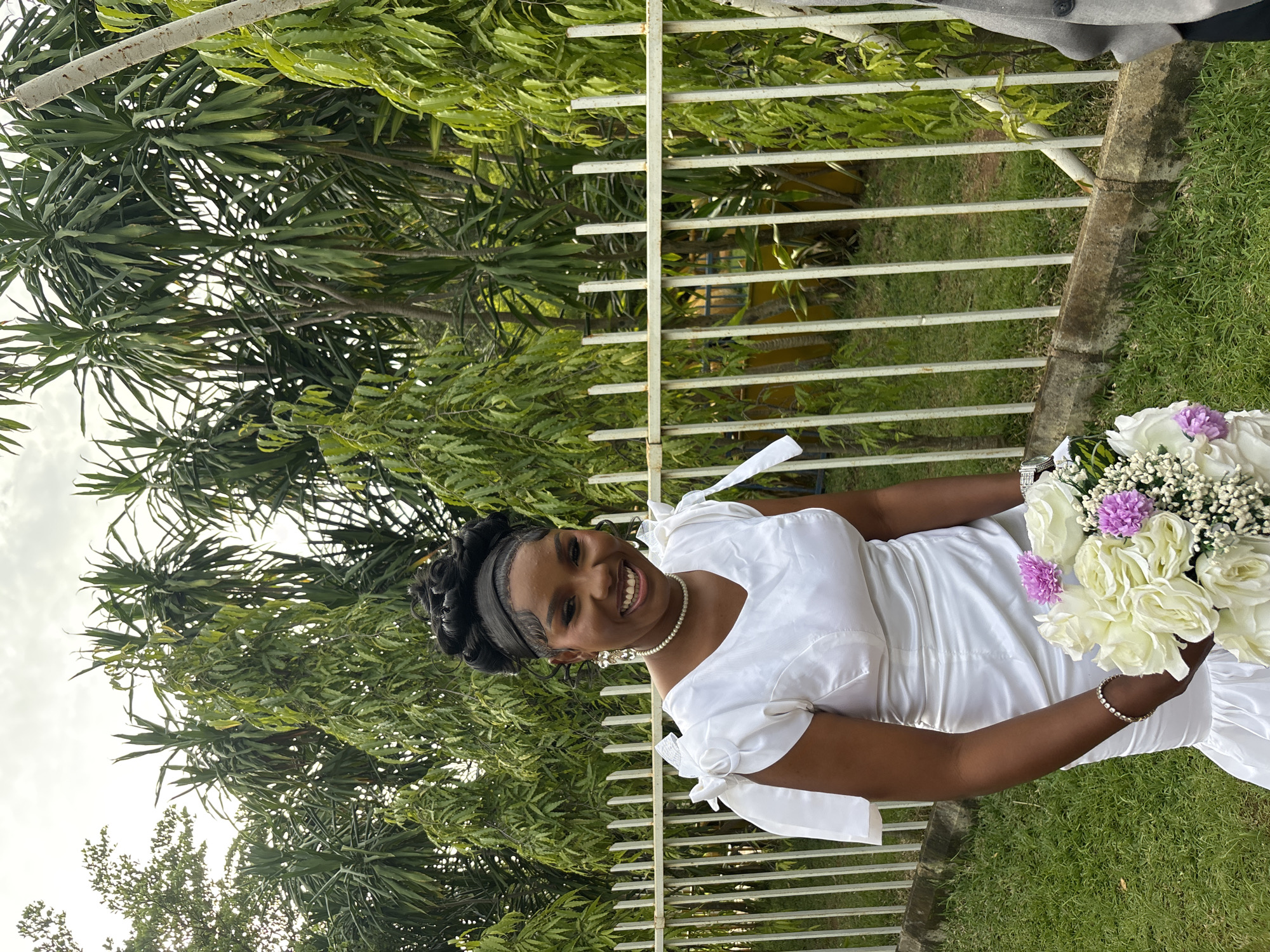 A young Black woman, adorned in a white dress with puffy sleeves, is captured in a portrait while holding a bouquet of white and purple flowers. She is smiling warmly, with her hair styled in an updo. The setting appears to be outdoors, with lush green foliage and a white metal fence visible in the background. The ground is covered in green grass, and a concrete border is present. The lighting suggests it might be daytime, possibly afternoon, with bright, natural light. The overall mood of the image is joyful and celebratory, hinting at a special occasion such as a wedding or graduation, with the woman's attire and the flowers contributing to this festive atmosphere. The image is oriented vertically, with the woman positioned on the right side, holding the bouquet in her right hand. The background is slightly blurred, focusing attention on the subject. The location is identified as Bukuru, Nigeria. No visible text is present in the image.