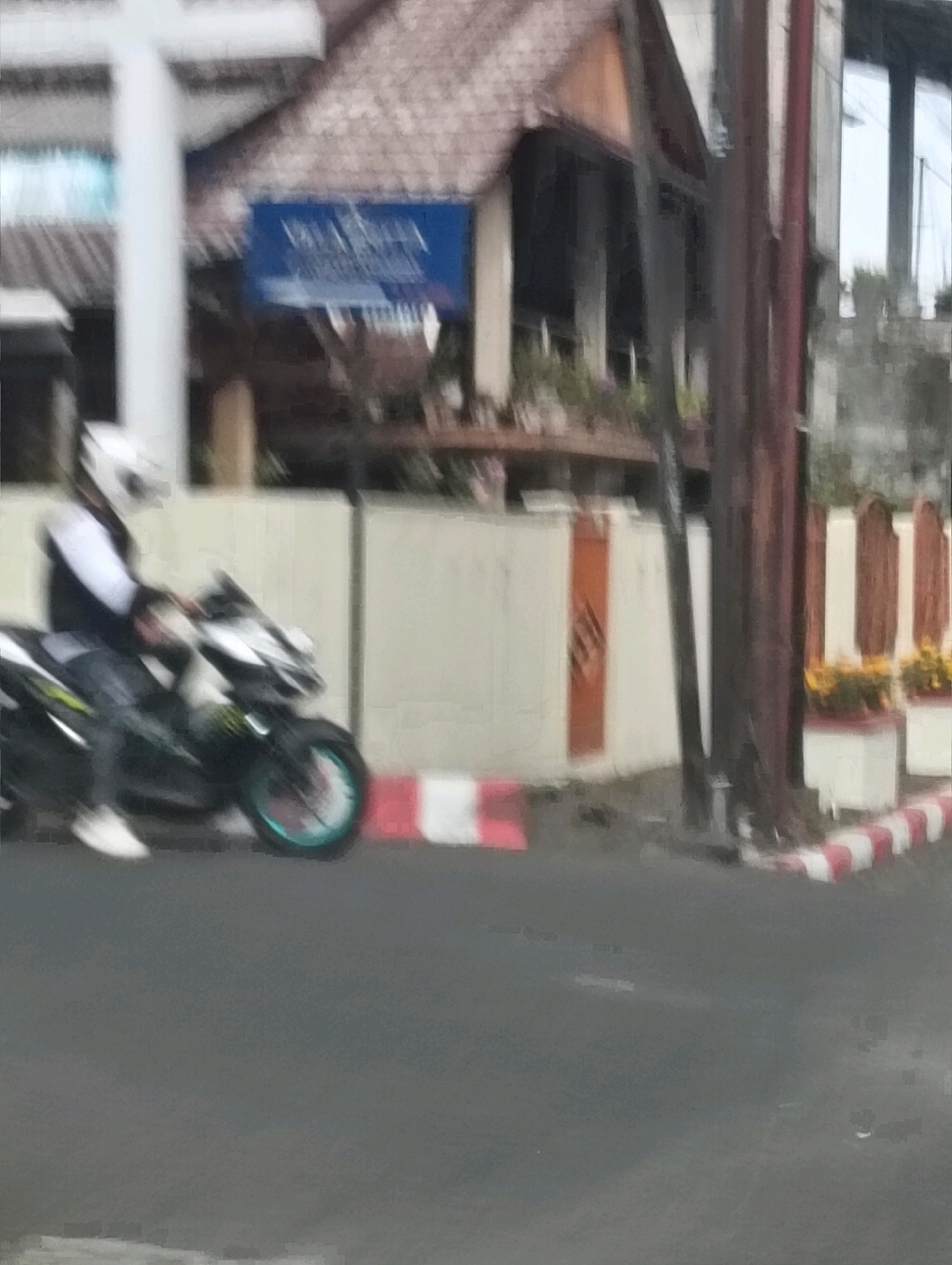 In Tomohon, Indonesia, a person wearing a white helmet is riding a motorcycle on a paved road. The motorcycle is dark colored with some green accents and has a teal rim on the front wheel. The rider is wearing a white shirt, a dark vest, and dark pants, with white shoes. The rider is in motion, as indicated by the blur effect on the motorcycle and the rider. Behind the rider, there is a light-colored wall with a red and white striped border. To the left of the wall, a white pillar is visible, and above it, a building with a thatched roof. A blue sign with white text is mounted on the building. The environment suggests an outdoor scene, possibly in a commercial or residential area.