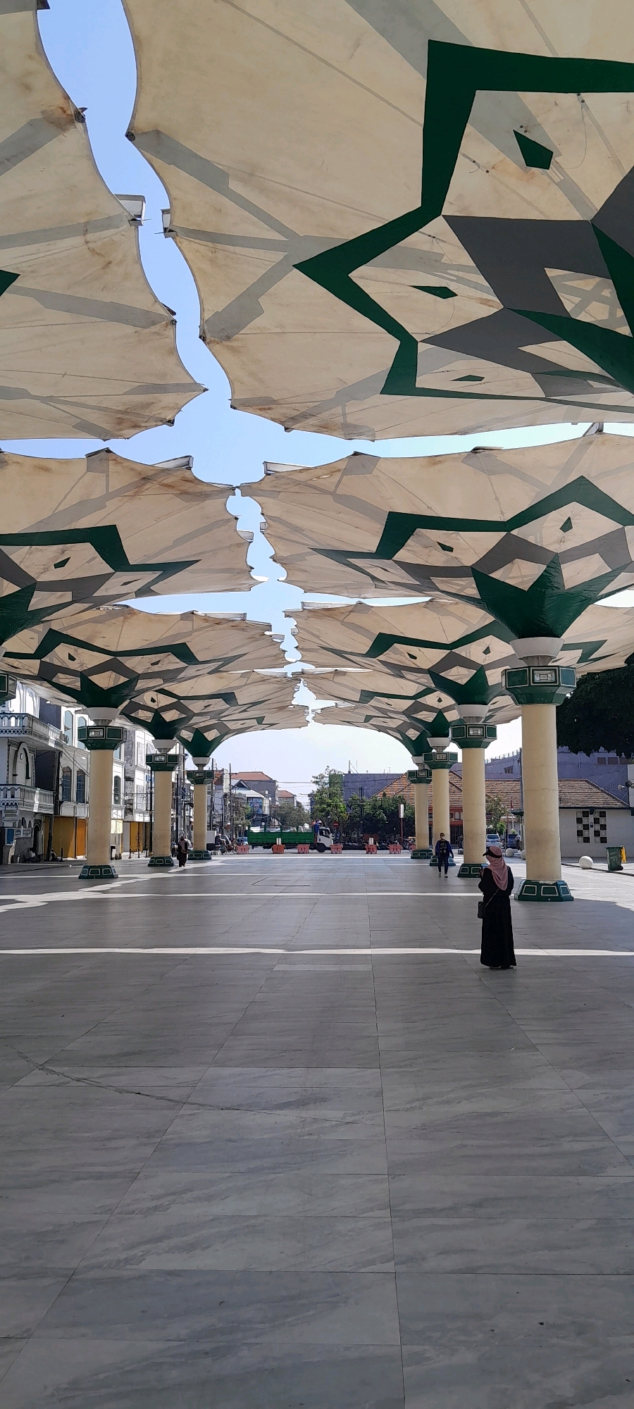 The image depicts an outdoor public space in the city of Pasuruan, Indonesia, likely a plaza or courtyard, covered by a series of large, white, umbrella-like canopies with green geometric accents. These canopies are supported by tall, cream-colored columns with green bases. The ground is paved with large, light gray tiles. In the foreground, the tiled ground is clearly visible, showing some wear and tire marks.
A woman wearing a dark abaya and a pink hijab is standing on the right side of the plaza, facing away from the camera, looking towards the open space. Her posture suggests she is standing still. Further in the background, on the left side of the plaza, a few other individuals are visible walking. The middle ground of the image shows a street with buildings lining it on the left and right sides, and a green truck with an orange barrier in front of it. The overall atmosphere is bright and sunny, with clear blue sky visible through the gaps in the canopies. The architecture suggests a location of cultural or religious significance.