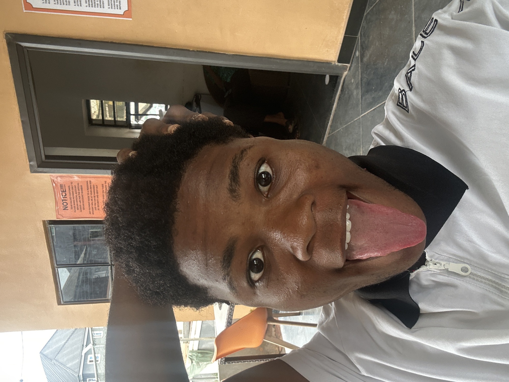 In Ihiagwa, Nigeria, a young person is taking a selfie outside. The subject is a Black male with dark, short, curly hair, wearing a white zip-up jacket with a black collar and a black emblem on the shoulder reading "BALEC". His right hand is raised to his head. He is sticking his tongue out at the camera, with his mouth open. In the background, there is an orange chair and a table, and a doorway leading into a building. There are two notices on the wall near the doorway. The overall environment appears to be an outdoor space attached to a building.