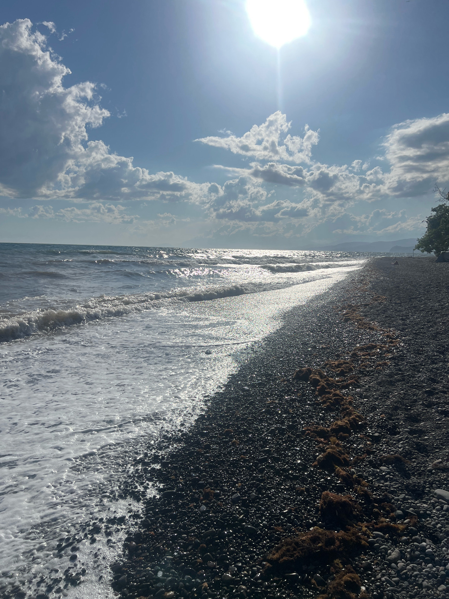 The image depicts a sunny day at a rocky beach. The sun is high in the sky, with bright rays shining down and reflecting off the water, creating a shimmering effect. The ocean waves are gently rolling onto the shore, with white foam washing over the dark pebbles. The beach is covered in smooth, dark rocks of various sizes, with some patches of brown seaweed scattered along the high tide line. In the distance, the coastline curves and mountains are visible on the horizon. The sky is blue with scattered white clouds. The overall scene is one of a peaceful, natural environment.