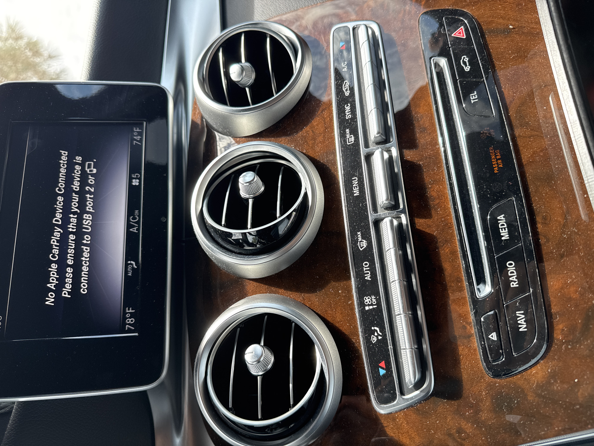 The image shows the interior of a car. In the center console, there are three circular air vents. Below the vents, there is a panel with buttons and a digital display. The display shows "No Apple CarPlay Device Connected Please ensure that your device is connected to USB port 2 or." Below this message, there are indicators for temperature (78°F and 74°F), A/C status (A/CON), and fan speed (5). The panel below the display has buttons labeled "MENU", "REAR", "SYNC", and "A/C". Further down, there is another panel with buttons labeled "NAVI", "RADIO", "MEDIA", "PASSENGER AIR BAG OFF", and "TEL". The car interior appears to have wood trim. The background is blurred, but appears to be outdoors, possibly a sunny day. The location is identified as Folsom, United States.