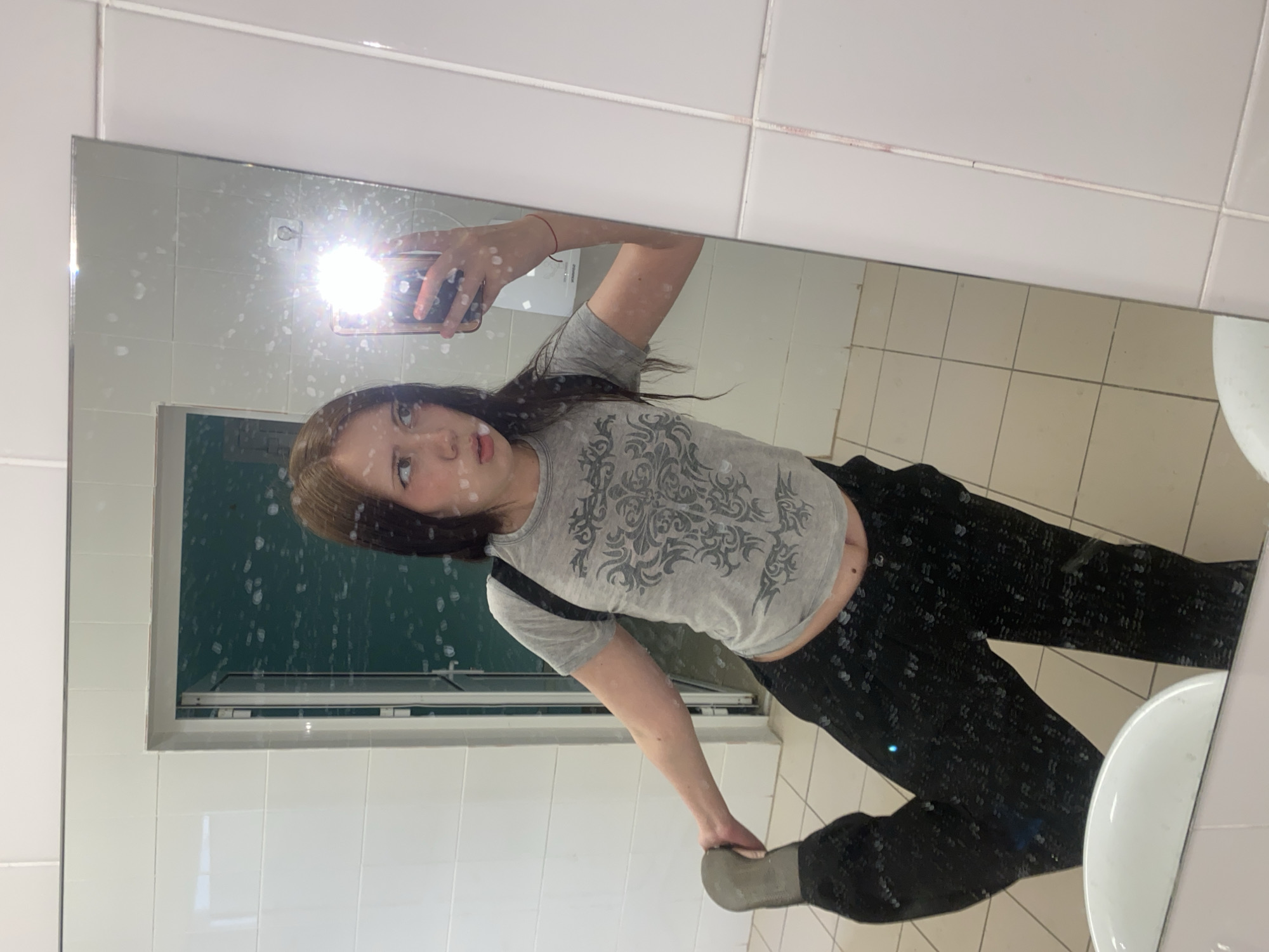 A young woman takes a selfie in a bathroom mirror. She is wearing a grey t-shirt with a patterned design and black pants. Her right foot is lifted as if she is posing or stretching. A sink is visible in the lower right corner of the reflection. The bathroom appears to be in a public facility, with white tiles on the walls and floor. A window or vent is behind her. The location is Unknown, Unknown.