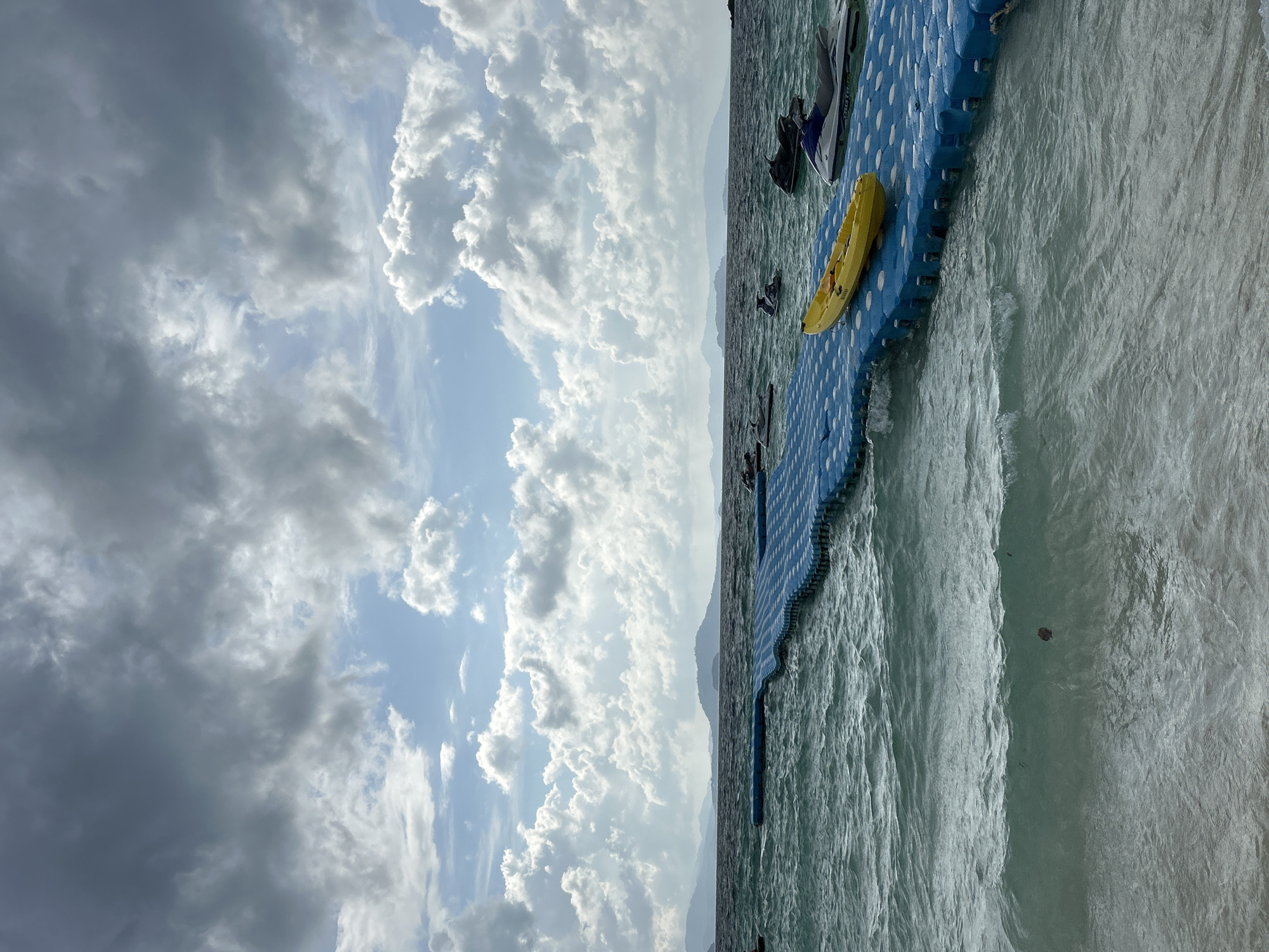 The image depicts a seascape with a blue modular floating dock extending from the shore into the choppy, turquoise water. On the dock, a bright yellow kayak is positioned in the foreground, and several jet skis are visible in the water surrounding the dock. In the distance, a hazy mountain range forms the horizon, beneath a dramatic sky filled with a mix of dark, heavy clouds and bright, fluffy white clouds. The scene is likely located near Phru Nai, Thailand, given the tropical appearance of the water, islands, and weather. The overall impression is one of a recreational area with water sports activities, under an overcast yet bright sky.