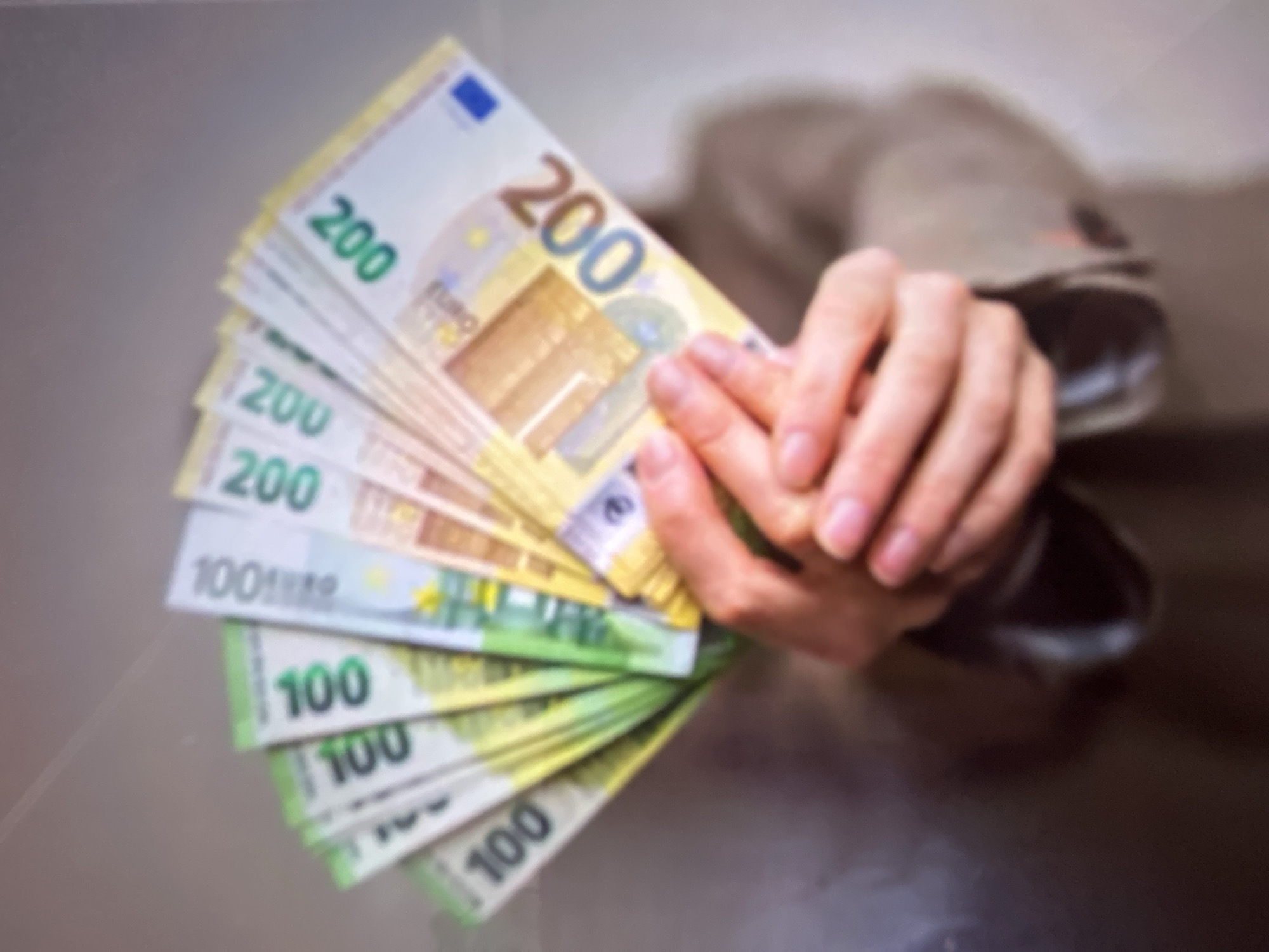 The image is a close-up, blurry photograph of a person's hands holding a fanned out stack of European banknotes. The denominations visible are 100 Euro notes and 200 Euro notes. The background is out of focus and appears to be a plain, light-colored wall. There is no indication of the city of Gelendzhik, Russia, or any other specific location. The person's attire is a light brown jacket or coat. The lighting is even and diffuse.