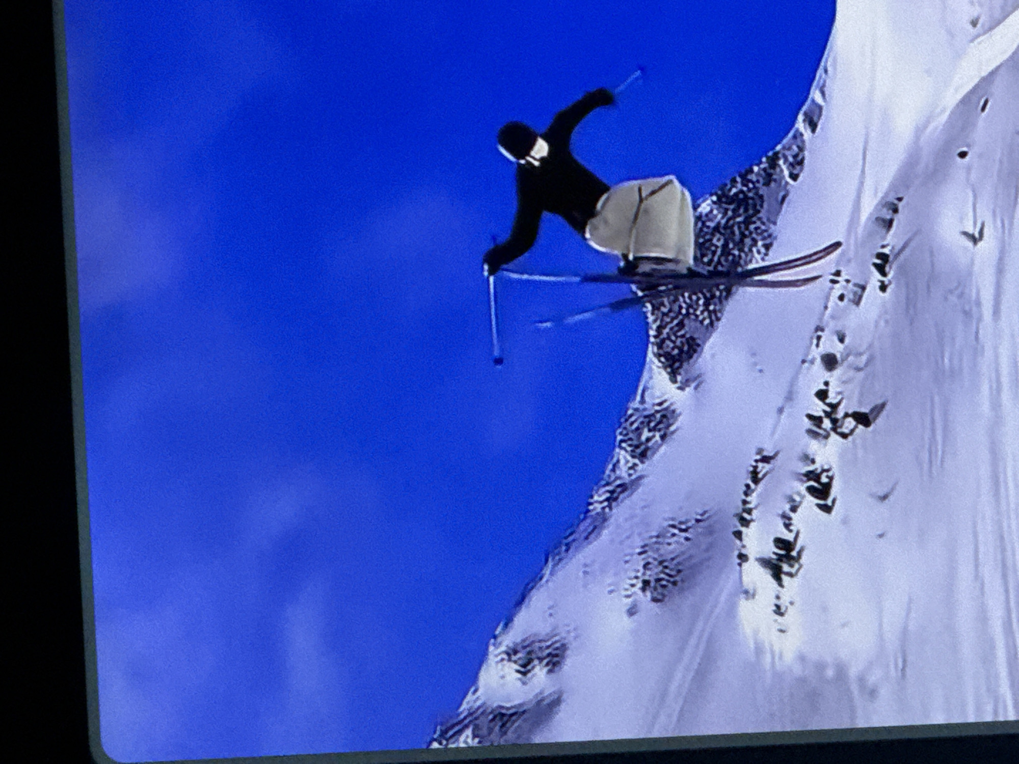 The image depicts a skier in mid-air performing a jump against a clear blue sky and a snow-covered mountain slope. The skier is wearing a black jacket, light-colored pants, and a black helmet with a white mask. They are holding ski poles in each hand. Their skis are visible, and they appear to be in a dynamic pose, suggesting an acrobatic maneuver. The mountain is covered in snow, with some dark rocky outcrops visible. The overall scene conveys a sense of extreme sport and athleticism in a natural, mountainous environment. There is no discernible location in the city of Cahul, Moldova, as the image portrays a mountain landscape.