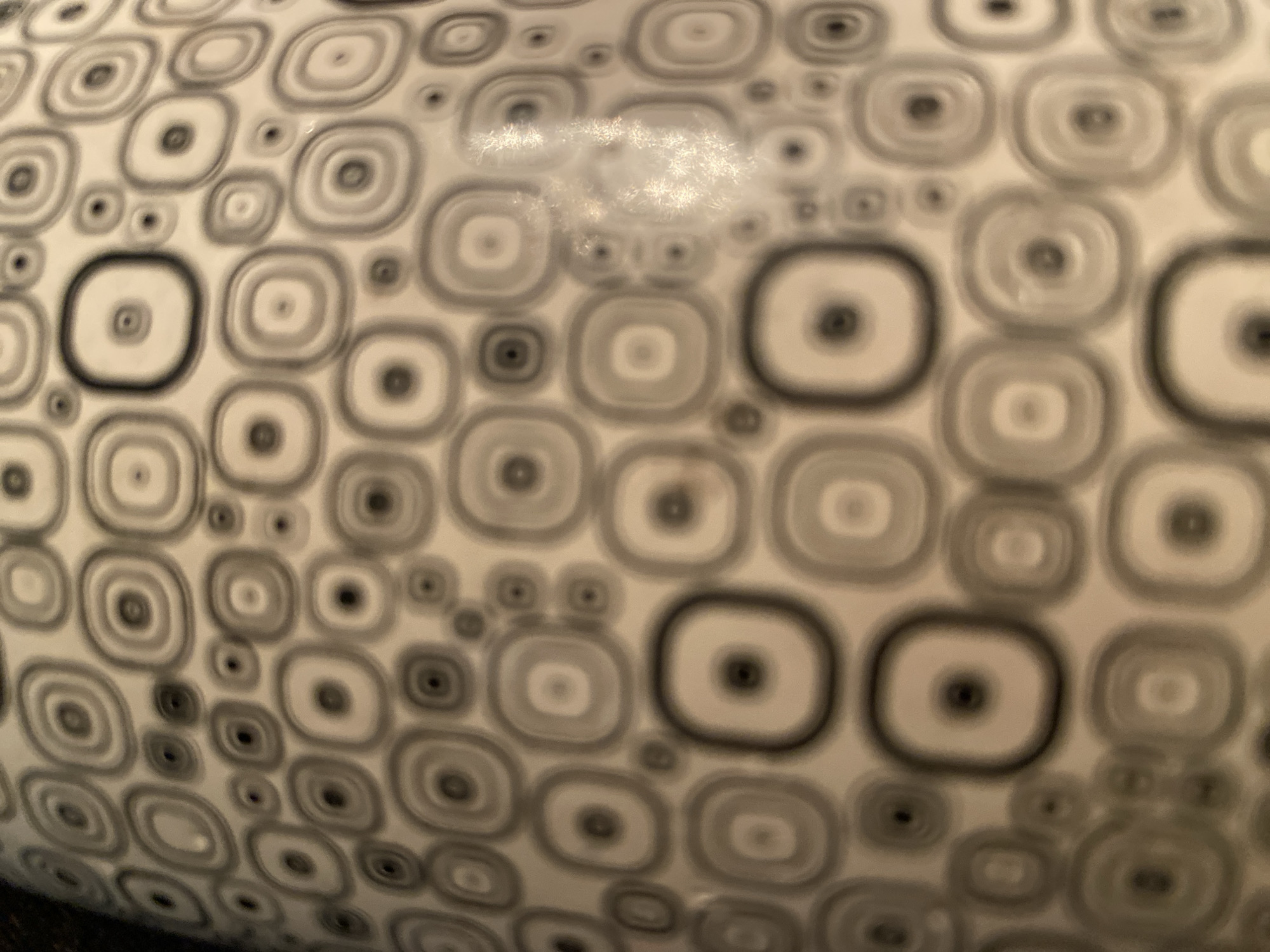 The image displays a close-up view of a white surface adorned with a repeating pattern of concentric circles and rounded squares in varying shades of gray and black. The pattern is dense and covers the entire visible area of the object, which appears to be cylindrical or spherical, given the curved perspective. There are no people, animals, or discernible landmarks visible in the image. The lighting suggests an indoor environment, with some glare reflecting off the surface, particularly in the upper right quadrant. The image does not contain any information that would allow for its localization in Ivanteyevka, Russia, or any other specific city.