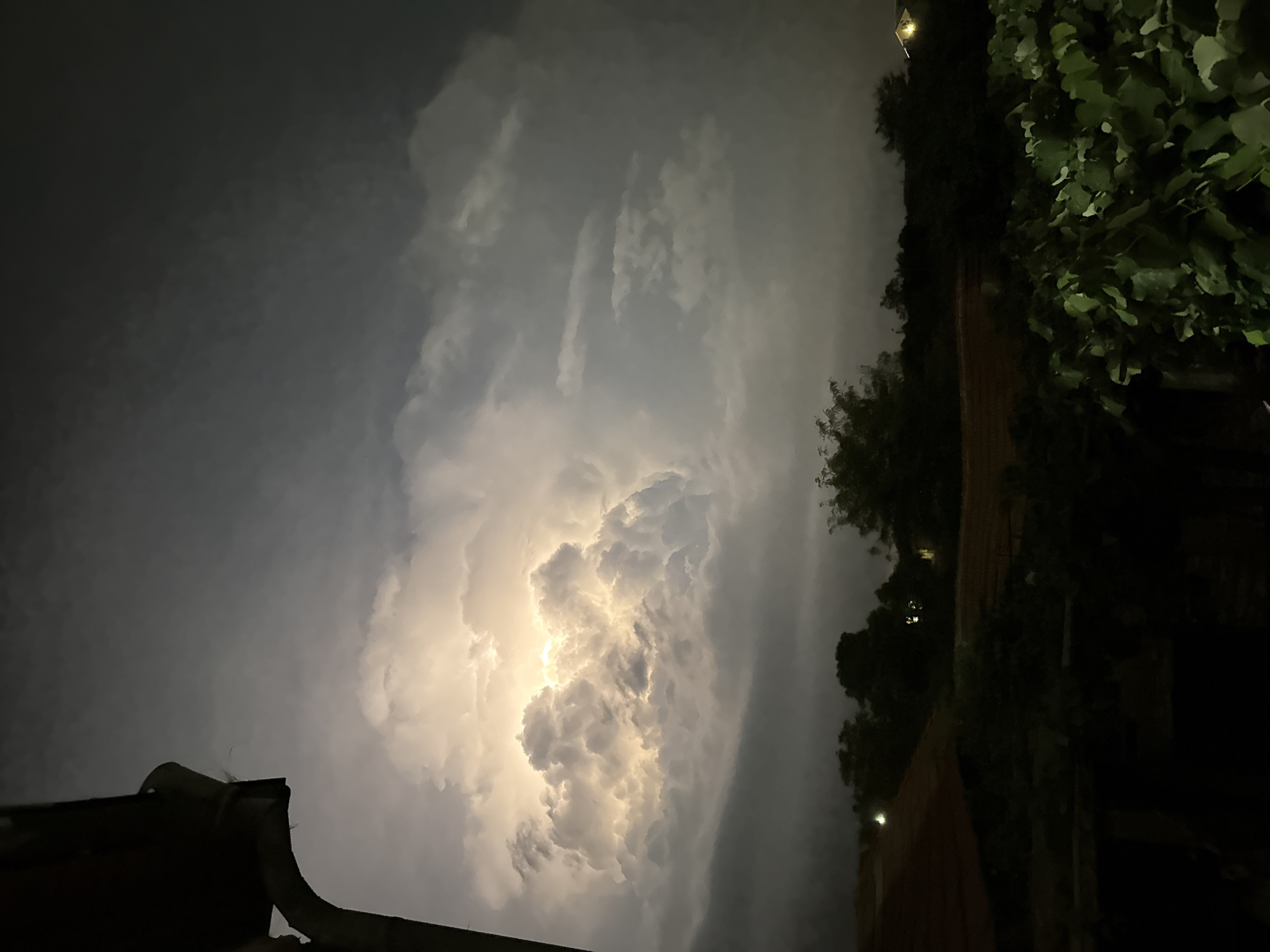 The image depicts a dramatic nighttime sky illuminated by a powerful lightning strike within a cumulonimbus cloud. The foreground features the dark silhouette of a building's gutter and downspout on the left side, casting a shadow. Below this, the tops of trees and vegetation are visible, with hints of a roofline and scattered lights suggesting a rural or suburban environment. The sky itself is dominated by the intensely bright, white-yellow flash of lightning, which brilliantly lights up the textured, turbulent clouds. The surrounding sky is a dark, moody gray, with other clouds creating a sense of depth and atmosphere. The location is the village of Ekzarh Antimovo, Bulgaria, on a stormy night.