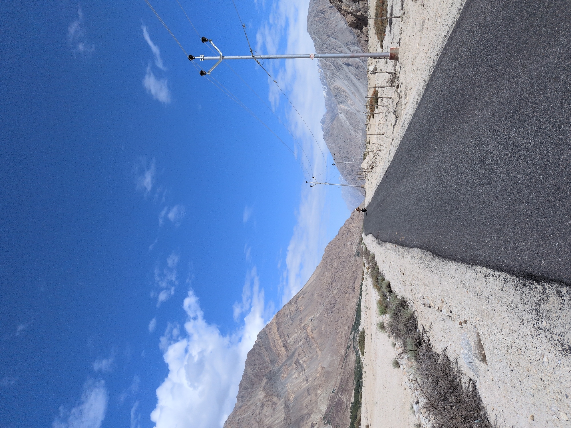 The image depicts a paved road winding through a dry, mountainous landscape under a bright blue sky with scattered white clouds. On the left, a large, layered mountain with rough, arid terrain dominates the background. The roadside is sandy and sparsely vegetated with dry bushes and scattered rocks. On the right side of the road, a telephone pole with multiple wires stretches across the frame, leading towards distant, rugged mountains. Further down the road, another telephone pole is visible. A single motorcyclist is seen riding away from the viewer on the road. The environment appears to be a remote, high-altitude region in India, potentially Sasoma, known for its challenging terrain and stark beauty.