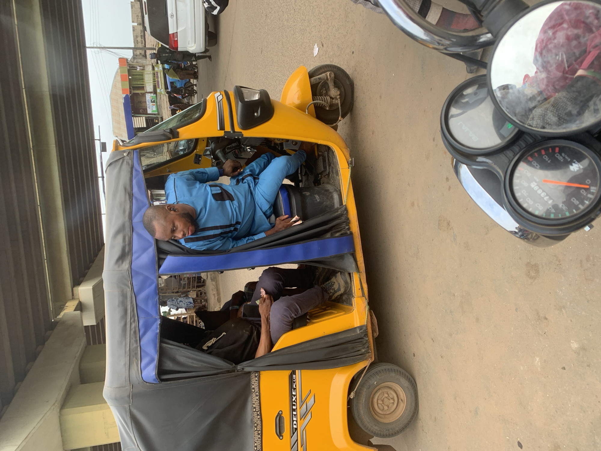 A man in a blue dashiki is seated in a yellow Bajaj auto-rickshaw, which is partially covered by a black and blue canopy. He is looking to his right and appears to be interacting with someone out of frame. Another person, wearing a black hoodie and dark pants, is seated to his left. The auto-rickshaw is on a dusty, unpaved road. In the background, there is a white SUV, other vehicles, and buildings with colorful awnings. An overhead bridge spans across the scene, casting shadows. The environment suggests a busy street in Shagamu, Nigeria, possibly near a market or transportation hub, given the presence of the auto-rickshaw and other vehicles typical of the area. The image appears to have been taken from the perspective of someone on another motorcycle, as indicated by the handlebars and dashboard in the foreground.