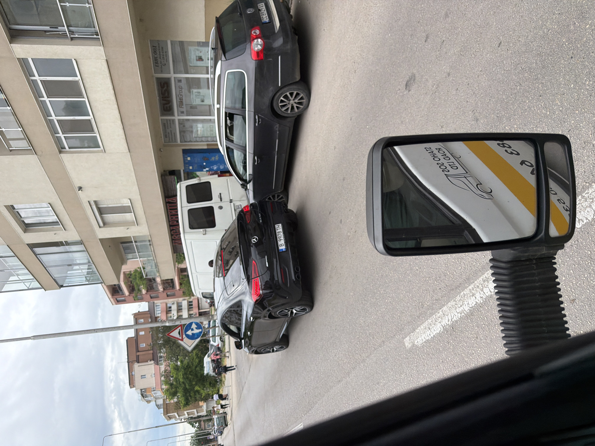 The image captures a street scene in Chervena voda, Bulgaria. A black Mercedes-Benz with license plate "B 9978 TH" and a dark-colored Volkswagen Passat with license plate "CC 8924 AX" are parked along the side of a building. The building features glass windows and a sign indicating "EVIS" and "FläktGroup." A white van is also present. A side mirror is visible in the foreground, reflecting a white vehicle with a yellow stripe and illegible text. A road sign with a pedestrian crossing symbol is also visible. People are walking in the background. The weather appears to be partly cloudy.
