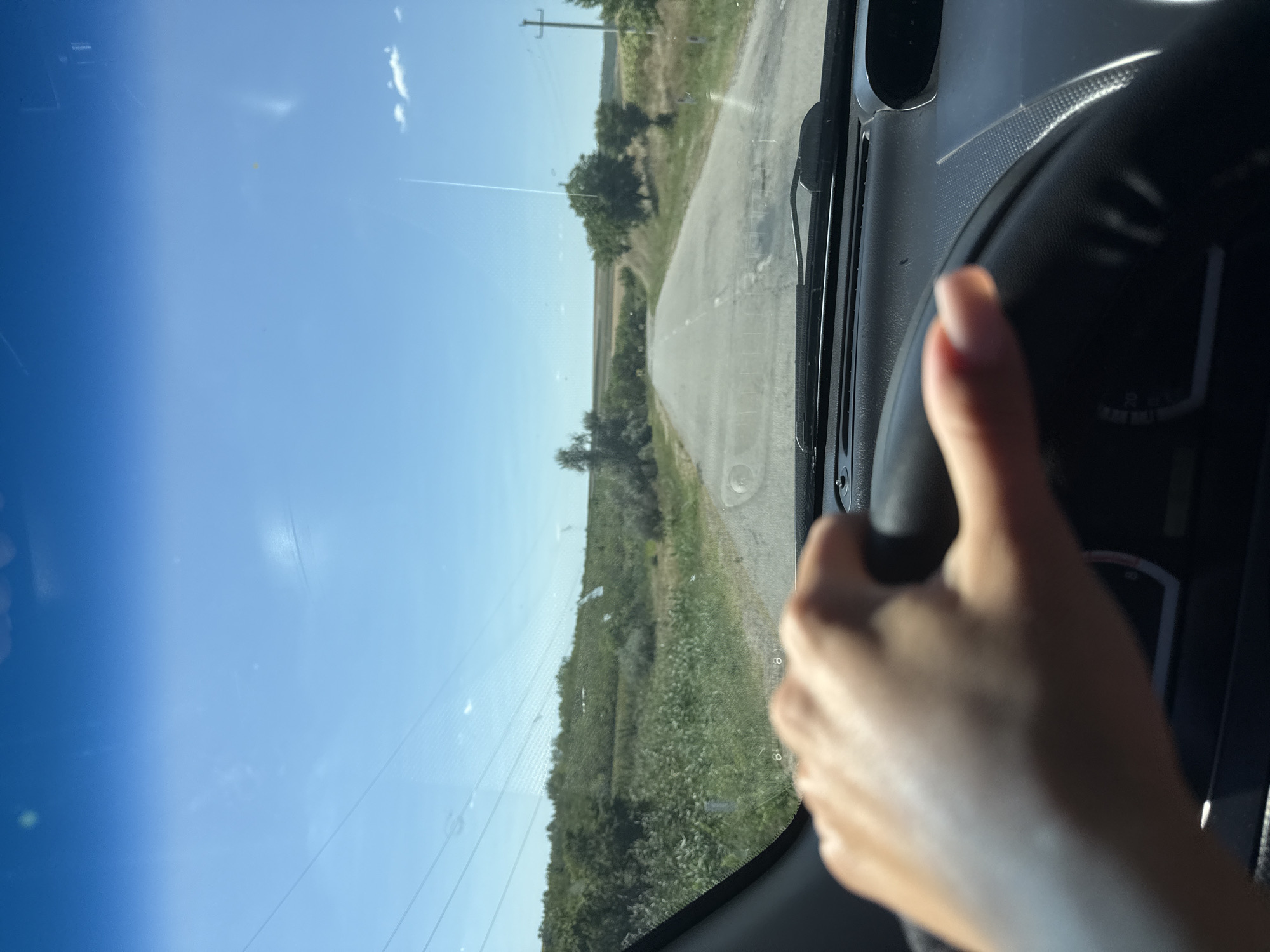 A person's hand grips a black steering wheel inside a car, driving down a paved road through a rural landscape under a clear blue sky. The car's dashboard is visible in the foreground, and the windshield shows a slightly smudged view of the road ahead, flanked by green fields and scattered trees. Power lines crisscross the sky. The scene suggests a sunny day in a rural area, likely on the outskirts of Basarabeasca, Moldova.
