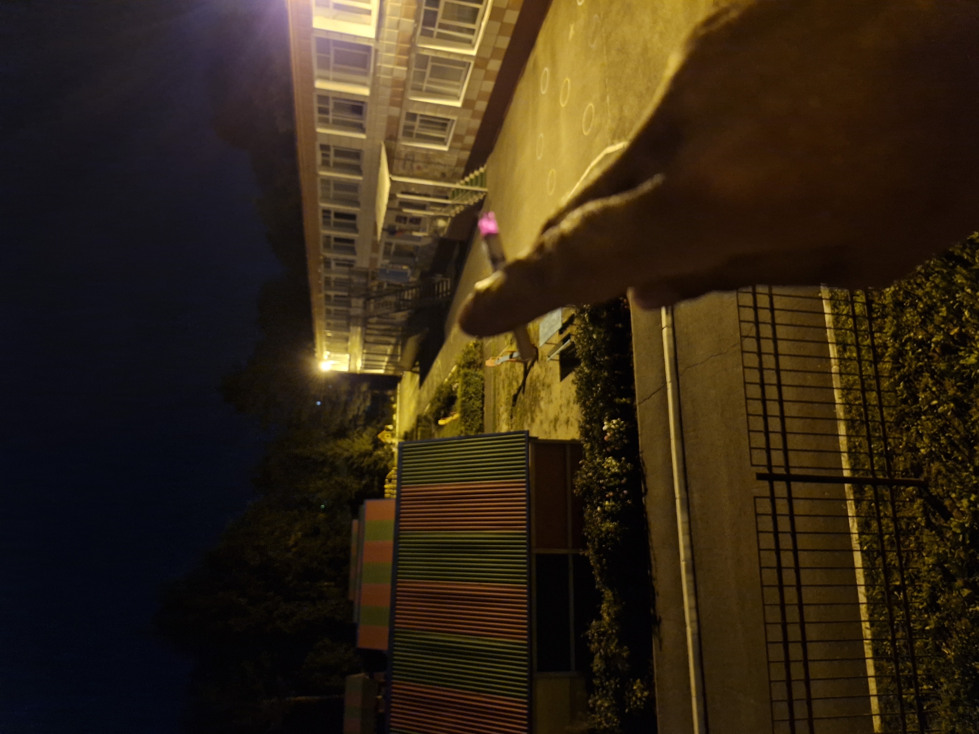 In the city of Kemerovo, Russia, at night, a person's hand is in the foreground, obscuring part of the view. The hand appears to be holding a cigarette that is lit, with a pinkish glow at the tip. In the middle ground, there are two colorful, striped structures, possibly children's playhouses or shelters, with red and green stripes. Behind these structures, a multi-story building with numerous windows and external staircases is illuminated by artificial light, suggesting it might be a school or residential building. The ground in front of the building is paved and marked with white circular patterns. A dark fence is visible in the foreground, separating a grassy area from a paved pathway or road. The overall environment suggests an outdoor area, possibly a courtyard or playground, illuminated at night.