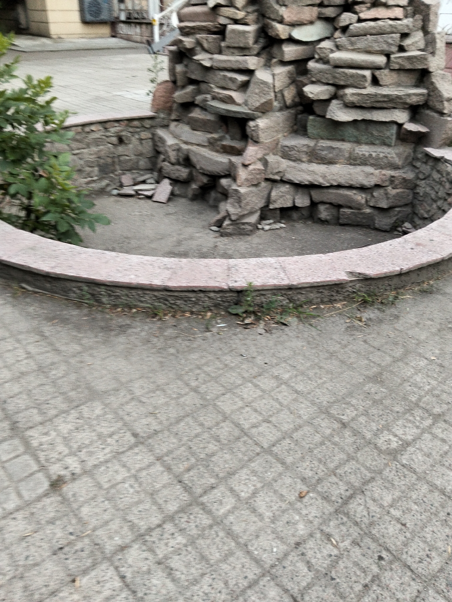 A low-angle outdoor photograph depicts a section of a stone structure and surrounding pavement in Almaty, Kazakhstan. The stone structure, built from rough-hewn, irregularly shaped rocks, rises in the background, featuring stacked layers and a stepped design. To the left of the structure, a green bush with broad leaves is partially visible. The foreground is dominated by a light-colored, patterned pavement, likely cobblestone, with a subtle grid of shadows cast across it. A low, curved wall with a pinkish-grey stone capping separates the pavement from the base of the stone structure. The ground within the curved wall appears to be dirt with some scattered debris. The scene is devoid of human presence or any discernible activity.