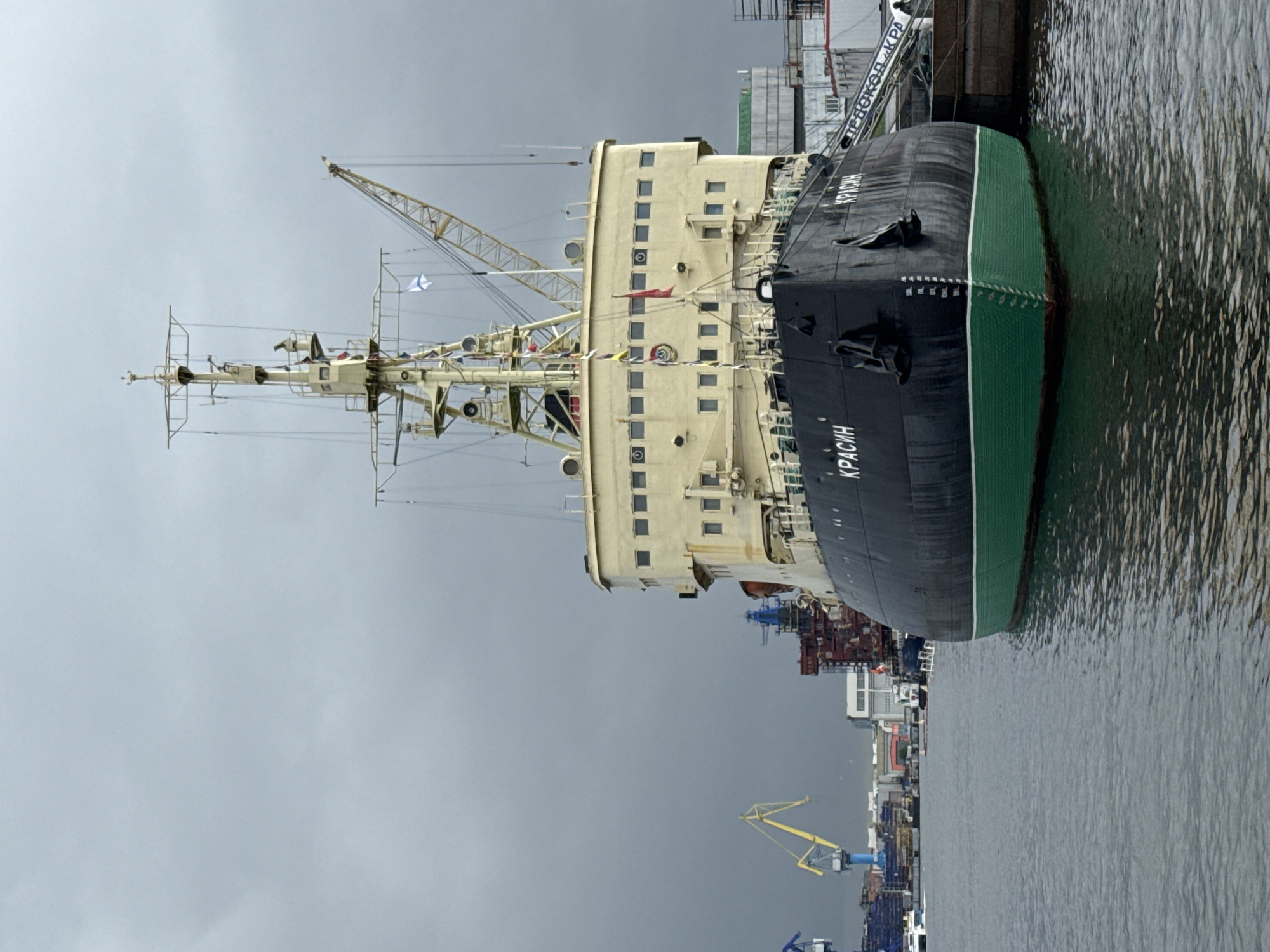 The image shows the icebreaker Krasin docked in an urban harbor under an overcast sky. The vessel has a black hull with green at the waterline, and the name "Krasin" is prominently displayed on the hull. A tall, multi-level cream-colored superstructure rises from the hull, with rows of windows visible. A crane structure is positioned on top of the vessel. In the background, there's a complex scene with harbor cranes, cargo containers, and other dockside structures. The water in the harbor has a rippled surface.