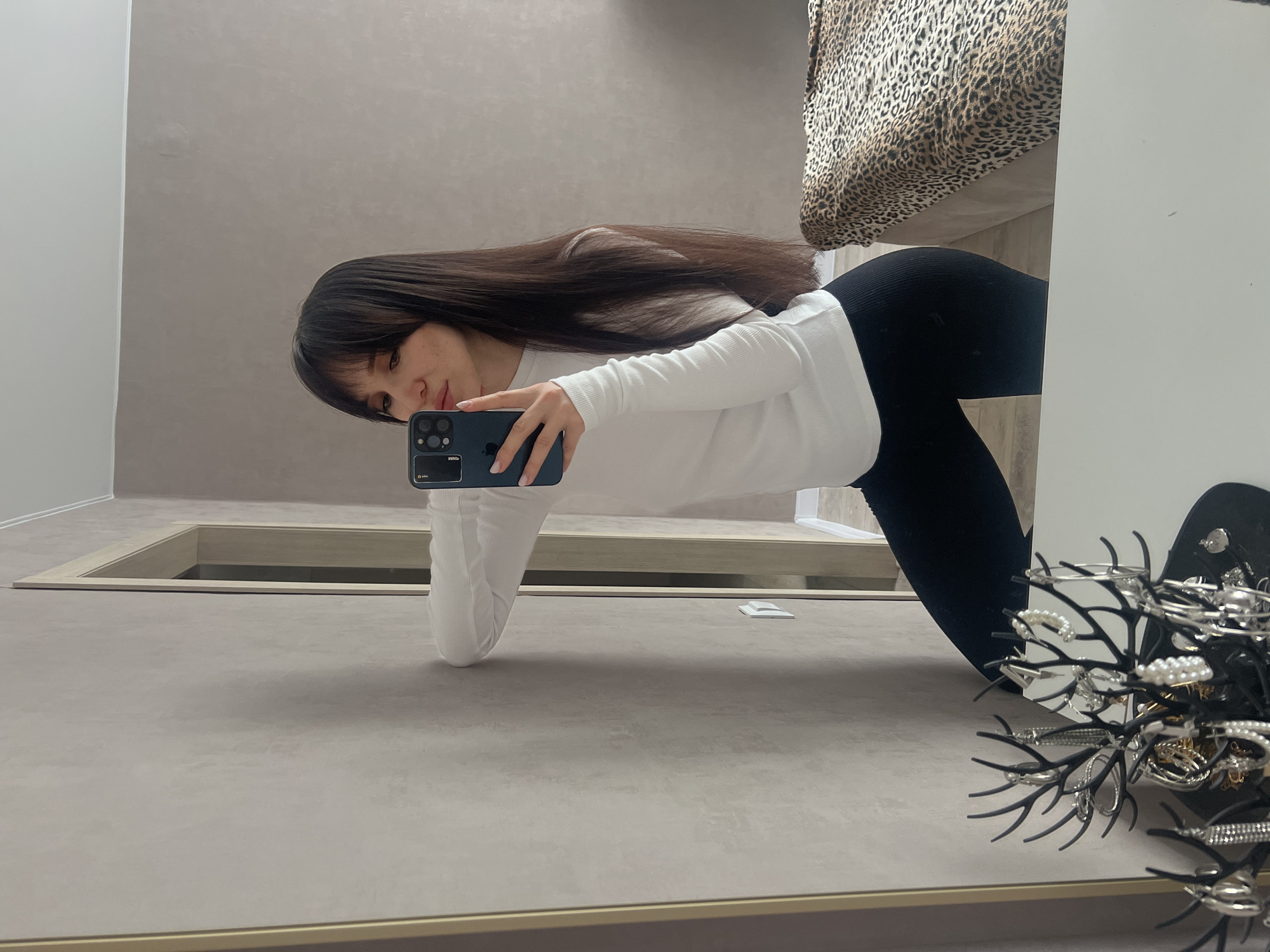 A young woman takes a photo of herself in a full-length mirror. She has long, dark hair with bangs and is wearing a white long-sleeved shirt and black leggings. She is holding a dark-colored smartphone up to take the photo.
The reflection in the mirror reveals a room with beige walls and what appears to be a recessed lighting fixture in the ceiling. There's also a glimpse of a bed with a leopard print covering. A black jewelry tree with various pieces of jewelry is located near the edge of the mirror.
The location is unknown, but assumed to be within the city of Votkinsk, Russia.
