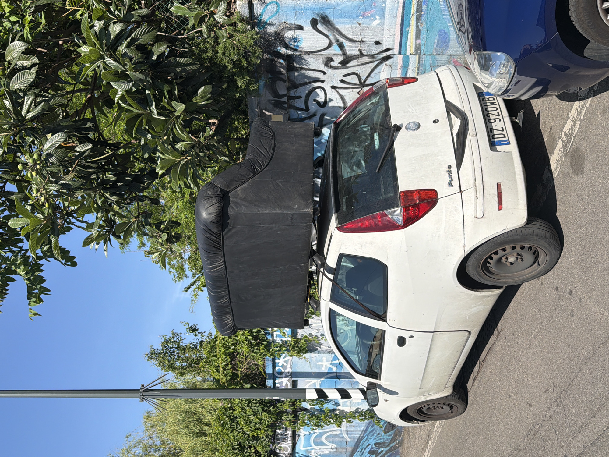 A white Fiat Punto is parked on a paved street, bearing the license plate "DZ-920WB," and transporting a black sofa on its roof. The vehicle is positioned next to a wall adorned with graffiti, with green foliage from nearby trees visible above and to the side. A blue car is partially visible to the right of the Fiat Punto. A light pole stands in the foreground, positioned between the sofa and the trees. The photo was taken in Milan, Italy.