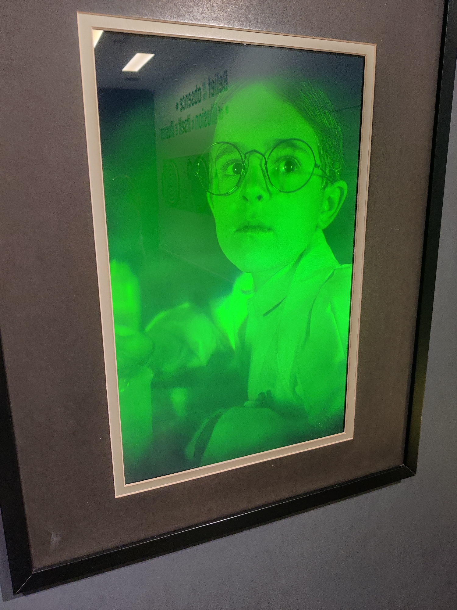 A framed portrait of a child wearing round glasses is displayed on a wall. The image is illuminated with a vibrant green light, giving it an otherworldly glow. The child's face is the primary focus, with their eyes open and looking directly at the viewer. They are wearing a collared shirt, and their hair is styled neatly. In the background, there are some blurry figures and what appears to be text on a wall, partially obscured by the green light. The frame is a double mat with a cream-colored inner mat and a dark outer mat, enclosed in a thin black frame. The portrait is hanging on a dark gray wall.