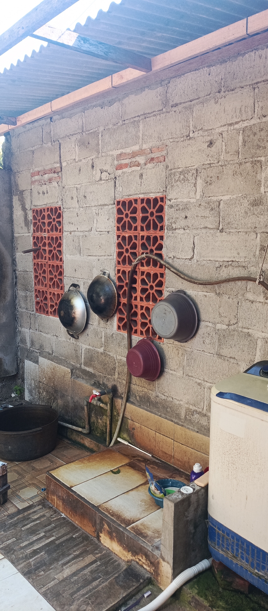 This image depicts an outdoor utility area, likely a washing area, located in Arjasa, Indonesia. The scene is characterized by a weathered brick wall with two decorative, orange-colored vent blocks on each side. Hanging from hooks on the wall are two dark-colored pots or pans and two plastic buckets, one maroon and one dark gray. Below the wall, there is a tiled basin with a red faucet and exposed plumbing. To the left of the basin is a large, dark, circular basin. To the right of the basin stands a white and blue washing machine, with a white hose extending from its base. The ground in front of the basin is paved with rough, dark-colored stones. The overall impression is one of a functional, somewhat utilitarian outdoor space.
