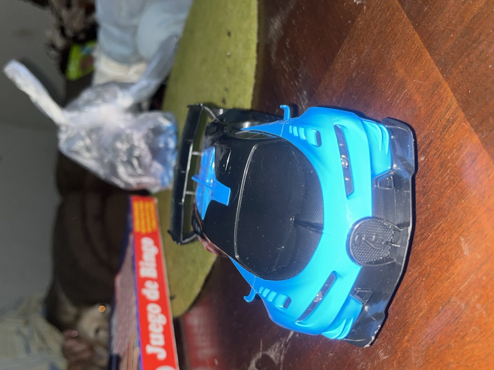 A blue and black toy race car is lying on a wooden table next to a "Juego de Bingo" box, partially obscured by a green object. A plastic bag containing an unidentifiable object is to the left of the "Juego de Bingo" box. The environment is indoors, with other indistinct objects visible in the background. The location is not specified in the image.