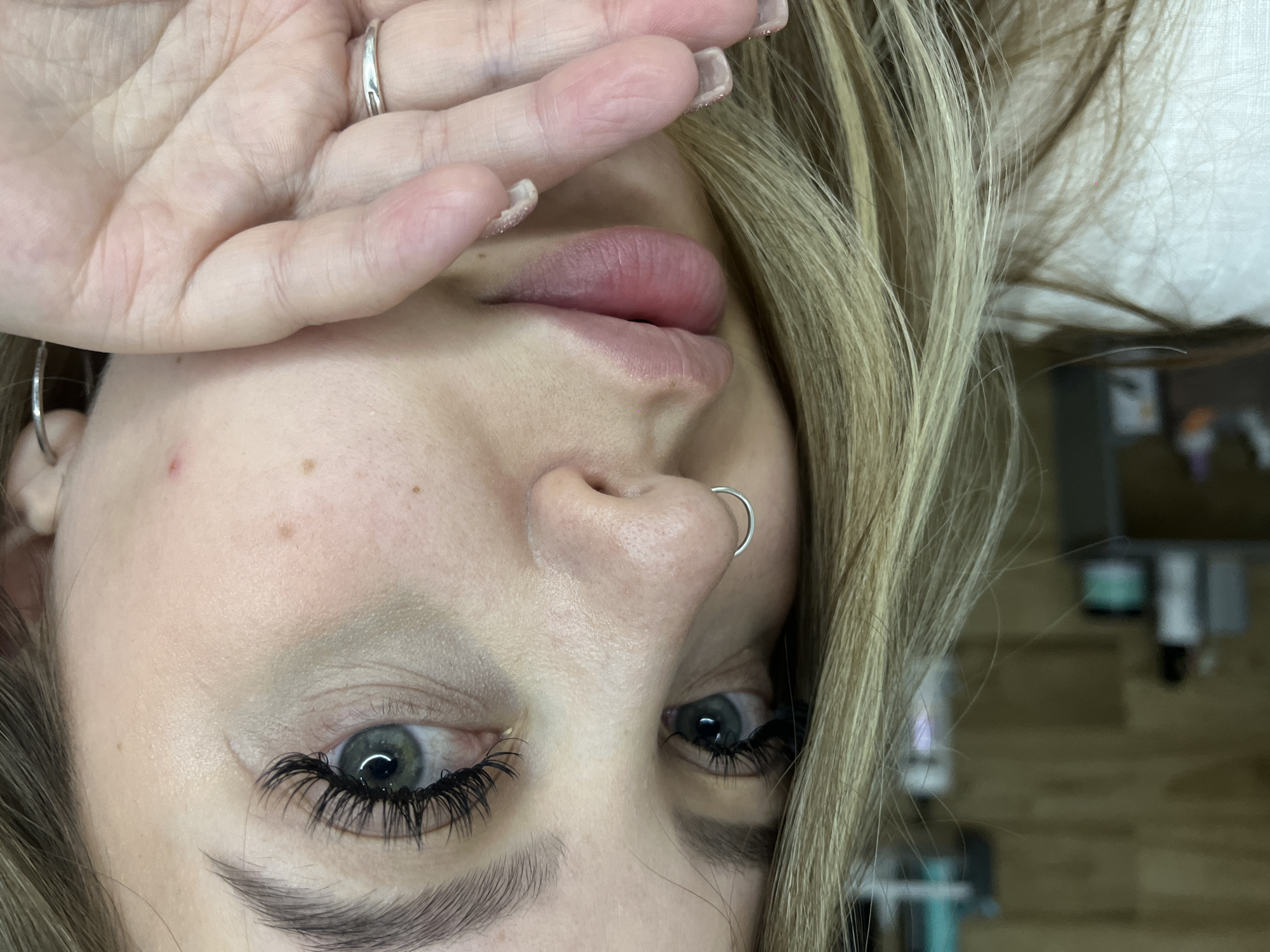 A close-up, portrait-style image features a young woman lying on her back, looking upwards. Her blonde hair frames her face, flowing outwards from her head. Her left hand is raised, partially covering her forehead and the upper part of her right eye. On her left ring finger, she wears a simple silver band. Her fingernails are visible, painted with a light, glittery polish.
The woman has fair skin, with a few small freckles or blemishes visible on her forehead and cheek. Both of her eyes are a striking light blue-green color, and she sports long, dark, voluminous eyelashes, likely extensions. Her lips are full and have a natural, soft pink hue. A silver hoop piercing adorns her septum. The background is heavily blurred, indicating an indoor setting with indistinct objects and shapes, possibly light-colored furniture or walls. There are no specific cues within the image to suggest a particular time of day or location, nor is any text visible. Her visible eye maintains a direct gaze towards the camera.