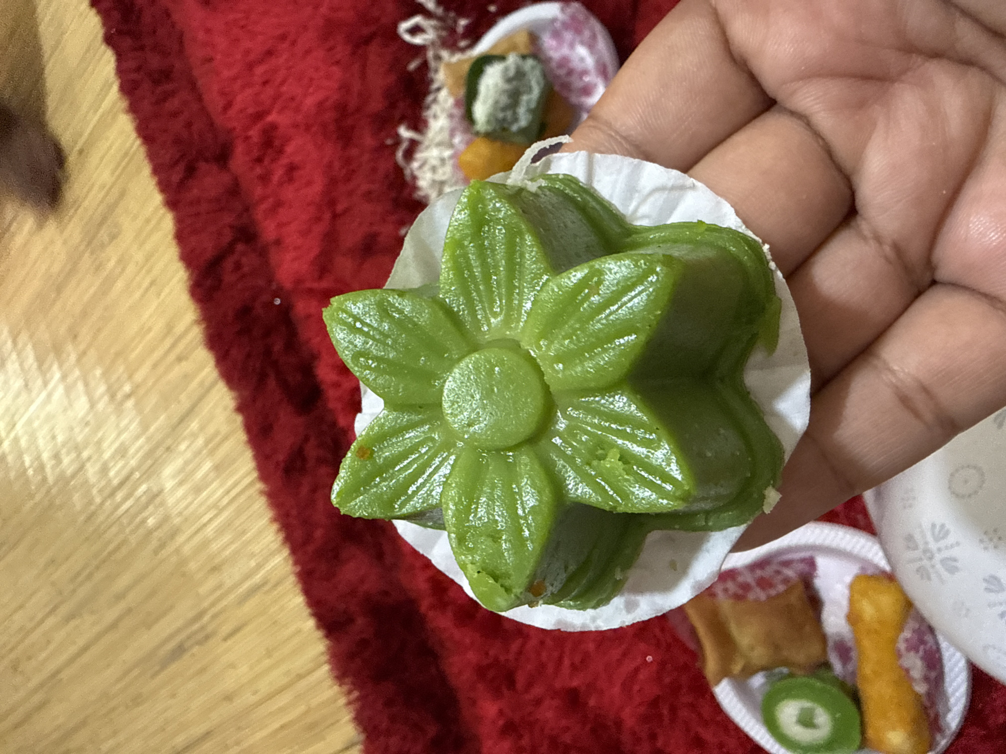 A hand holds a green, flower-shaped sweet in a white paper cup. Other sweets are on plates in the background. A red fabric and a light wood surface are visible. The location is not discernible.