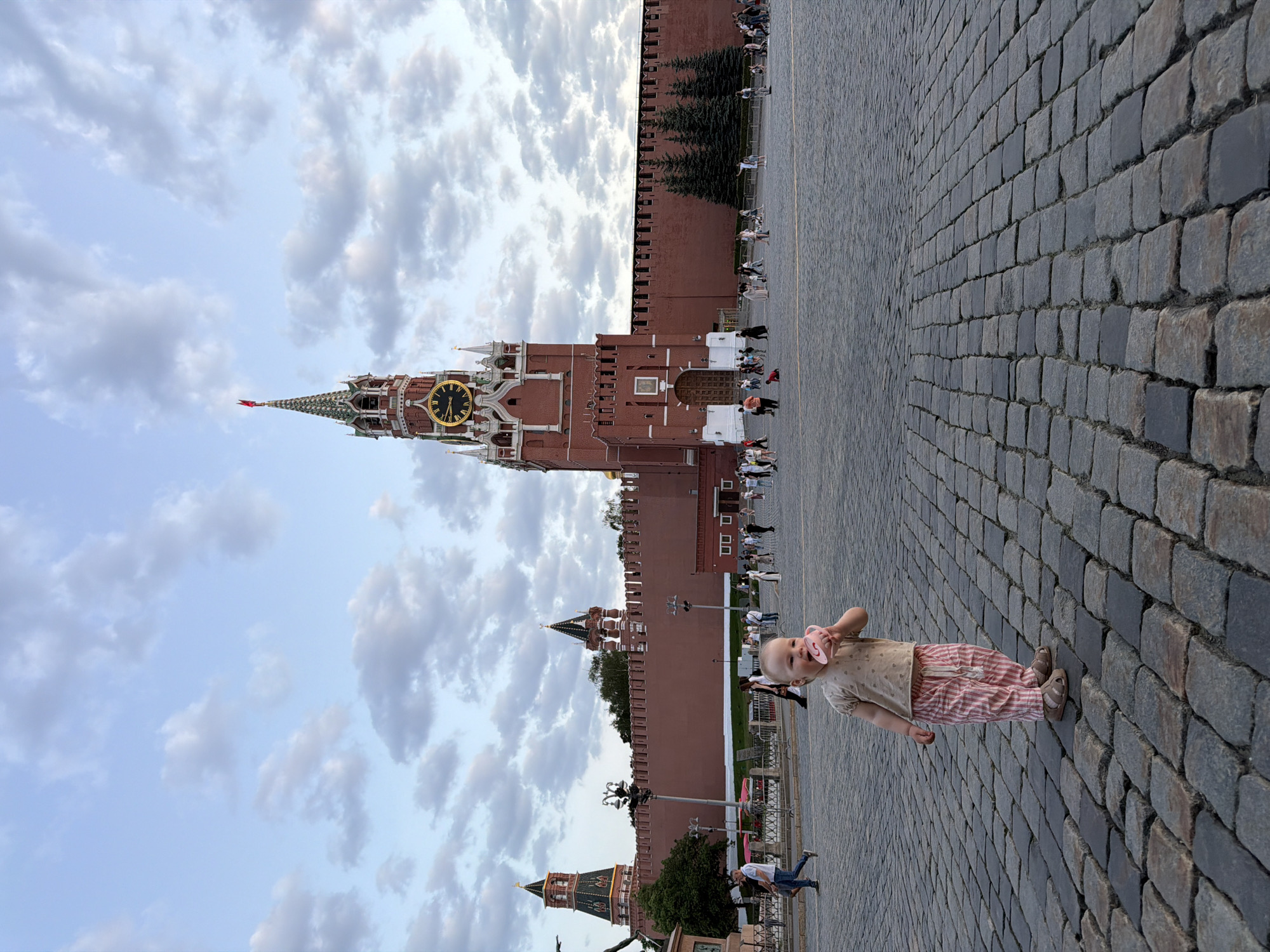 The image captures a low-angle shot of a child standing on a cobblestone pavement, with the Moscow Kremlin as a backdrop. The child, wearing a beige shirt and red-and-white striped pants, is facing the camera, holding a pink pacifier. The pavement is composed of grey cobblestones. Behind the child stands the Moscow Kremlin, a fortress complex with red brick walls and towers, including the Spasskaya Tower with its prominent clock. In the background, the sky is visible with a mixture of blue and grey clouds. Other people are walking or standing near the Kremlin walls. The image is taken in the Red Square of Moscow, Russia.