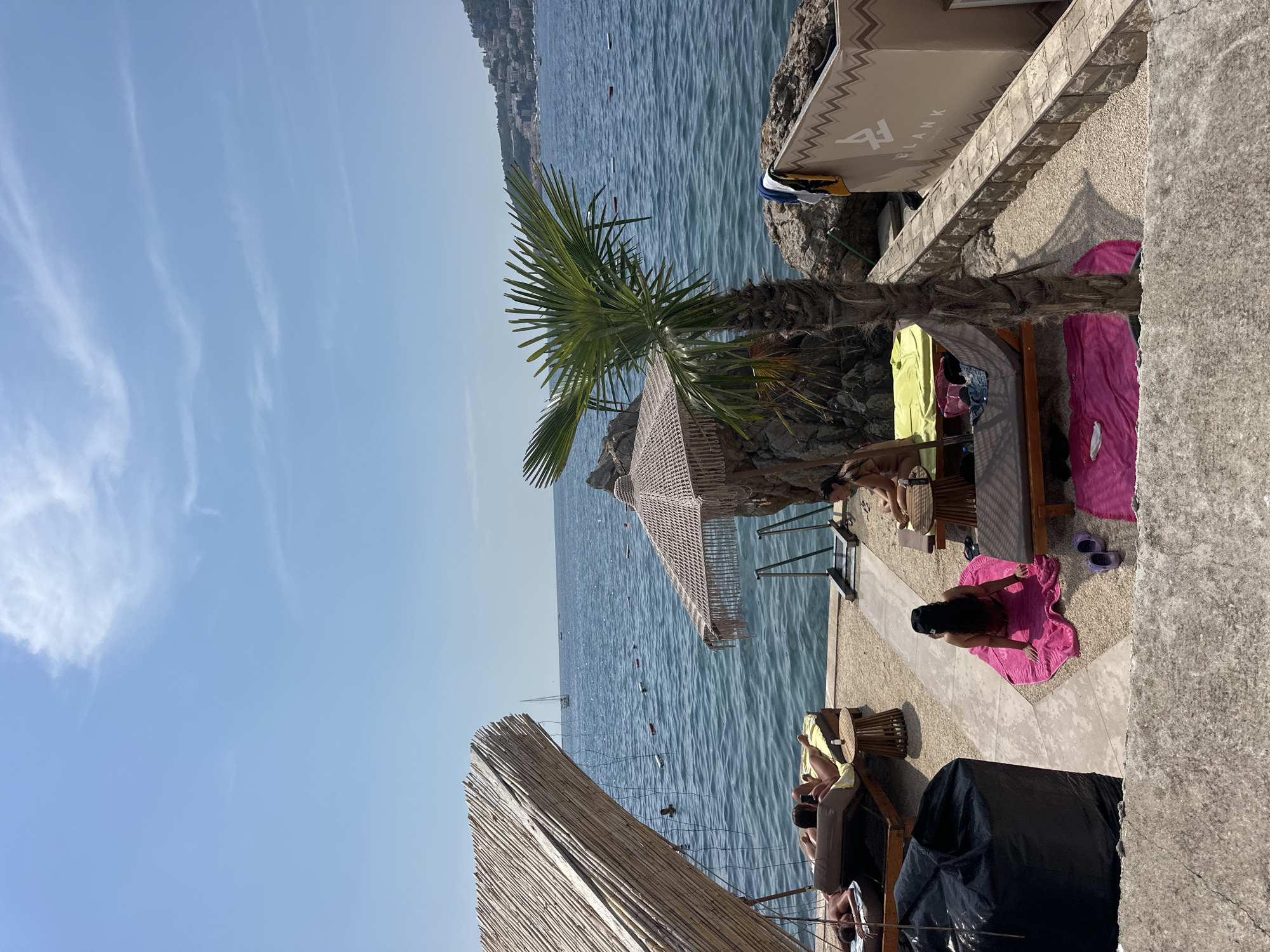 The image depicts a section of a beach resort in Budva, Montenegro, overlooking the Adriatic Sea. In the foreground, a rough concrete structure forms a ledge. Beyond this, a gravel and paved area serves as a lounging space. Several lounge chairs with towels are arranged, some occupied by sunbathers. A woman with dark hair, wearing a black baseball cap, is seated on a bright pink towel, facing away from the camera. To her left, another woman is seated on a lounge chair, also facing away. Further back, two people are lying on lounge chairs.
A prominent thatched roof structure with a bamboo-like texture is situated on the left side of the frame, providing shade. Adjacent to it, a palm tree stands tall, its fronds reaching towards the sky. Another shaded area with a macrame-style umbrella is visible, accompanied by metal railings leading towards the sea.
The sea is calm and blue, with several buoys marking a swimming area. A sailboat is visible in the distance. The coastline on the right side of the image shows a hilly landscape with buildings dotting the slopes. A branded structure with the text "BLANK" is present on the right side of the beach area. The sky is clear with a few wispy clouds.