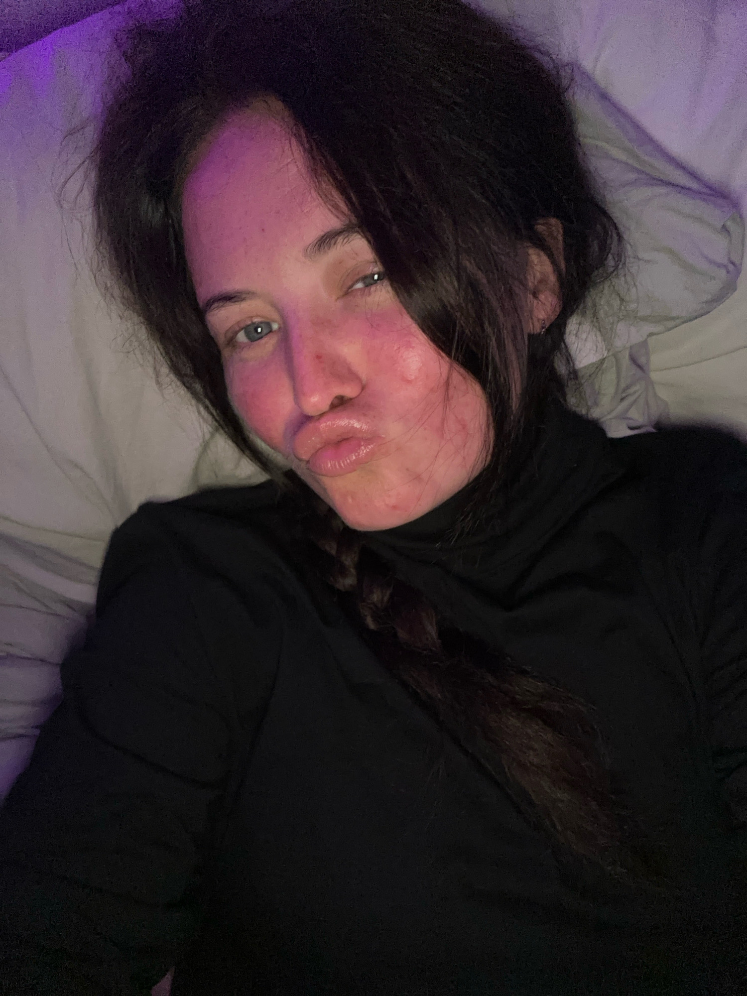 A young woman with dark hair is lying in bed, looking into the camera with a pout. She is wearing a black turtleneck. The room is lit with purple lighting, and the bedding is white. The woman's face is slightly flushed, and she has light blue eyes. Her hair is styled in a braid. There are no visible indications of the specific location within Günzburg, Germany.