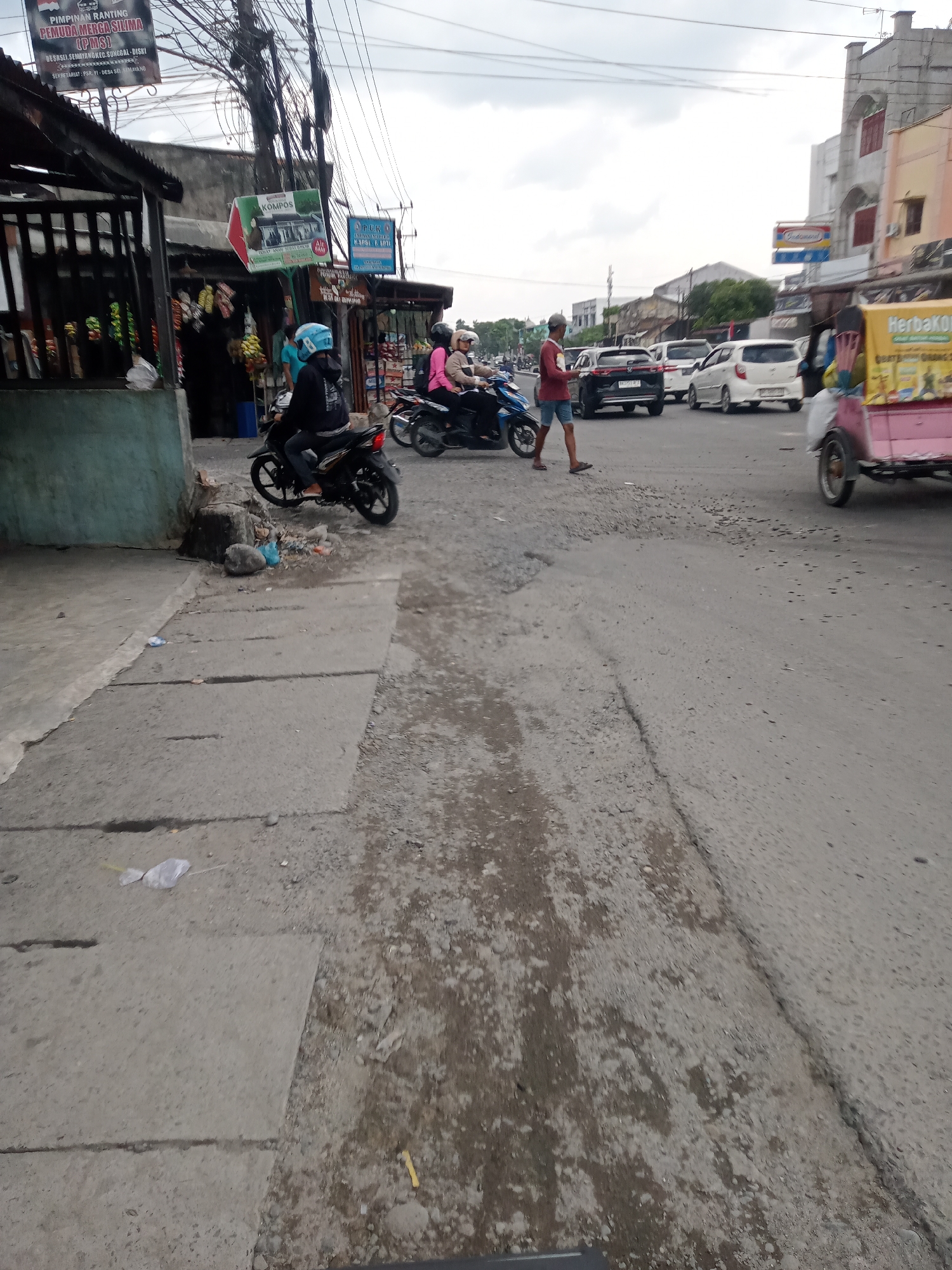 The image depicts a street scene in the city of Sumber Melati Diski, Indonesia. In the foreground, a person wearing a blue helmet rides a black motorcycle. To their right, two other individuals are on a motorcycle, one wearing a pink shirt and the other a black shirt. A pedestrian in a red shirt and blue shorts walks across the road. Several vehicles, including cars and motorcycles, are visible in the background, suggesting moderate traffic. The road surface appears to be asphalt with some areas of wear and tear, including potholes and uneven patches. Along the side of the road, there are shops with awnings and various items displayed outside. Overhead, a dense network of electrical wires crisscrosses the sky. The overall atmosphere is that of a bustling urban environment, possibly during the daytime, with overcast weather conditions.