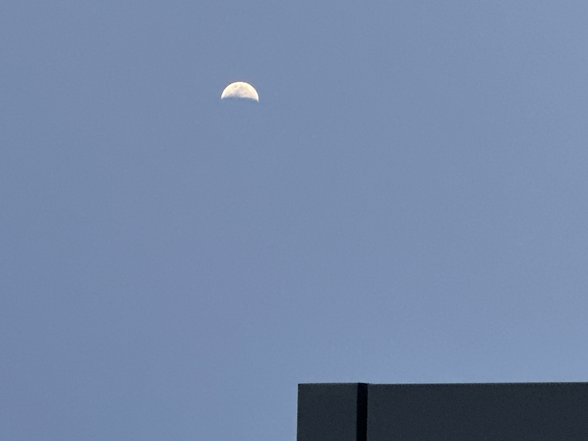 The image shows a half moon in a blue sky, positioned above a gray, blocky structure. The structure appears to be a building or a part of a building, with distinct edges and a vertical division. The image suggests an urban setting, although specific location details like the city of Chaikovsky, Russia, cannot be discerned from the visual content alone. The scene is devoid of people or other obvious signs of activity.