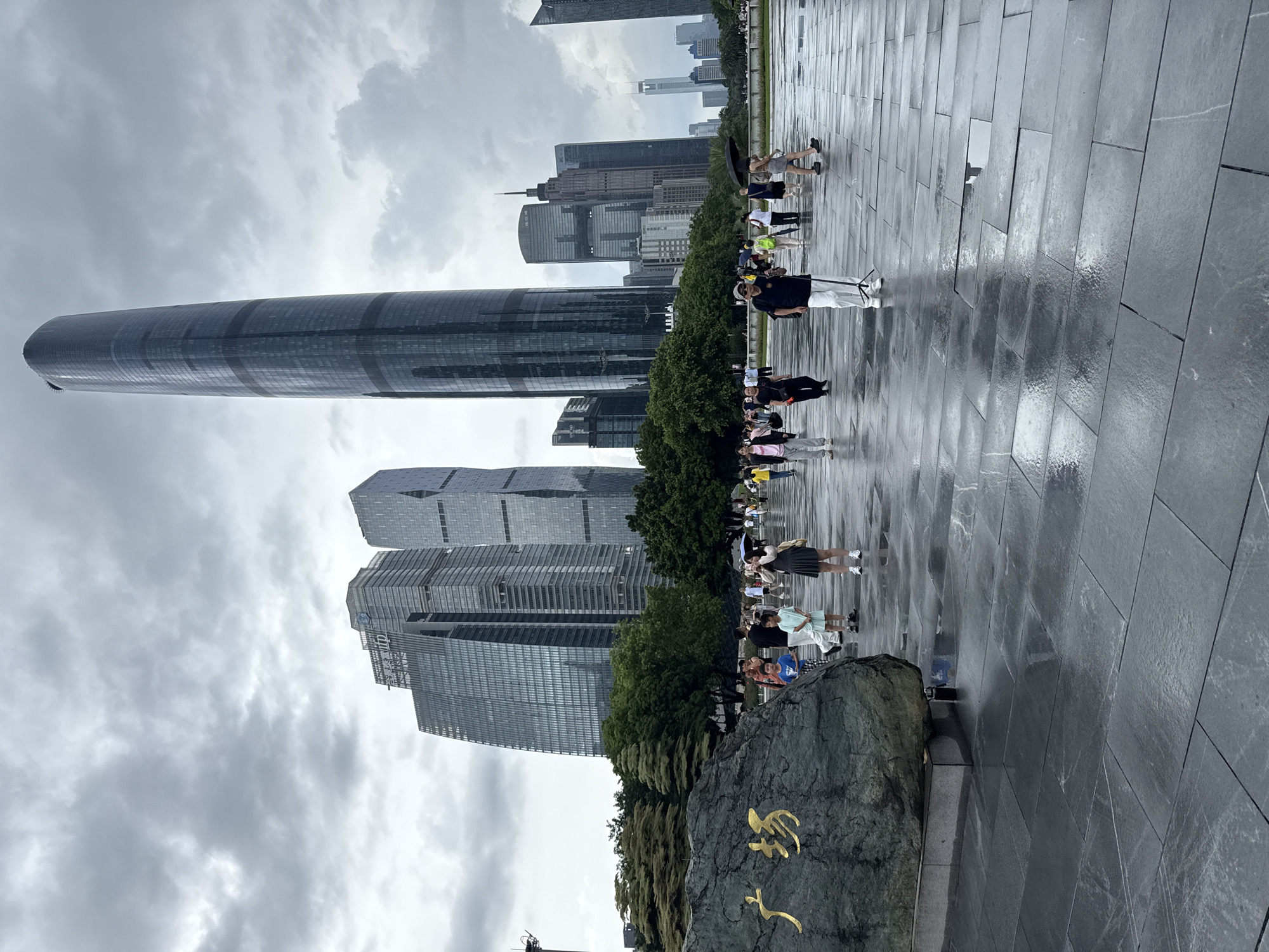 This image depicts the Tianhe District in Guangzhou, China, a modern urban landscape characterized by impressive skyscrapers. In the foreground, a large, dark rock with golden Chinese characters is prominently placed, possibly a landmark or monument. The ground, paved with large, dark tiles, is wet and reflective, indicating recent rain or damp conditions. Numerous people are scattered across the plaza, some walking individually, others in small groups. They are dressed in casual attire, and a few are holding umbrellas. The sky is overcast and cloudy, contributing to a somber atmosphere. The background is dominated by a cluster of tall, modern buildings, including a particularly distinctive, slender skyscraper with a curved top. Lush green trees are visible between the buildings and the foreground. The overall scene captures a moment in an urban public space under a cloudy sky.