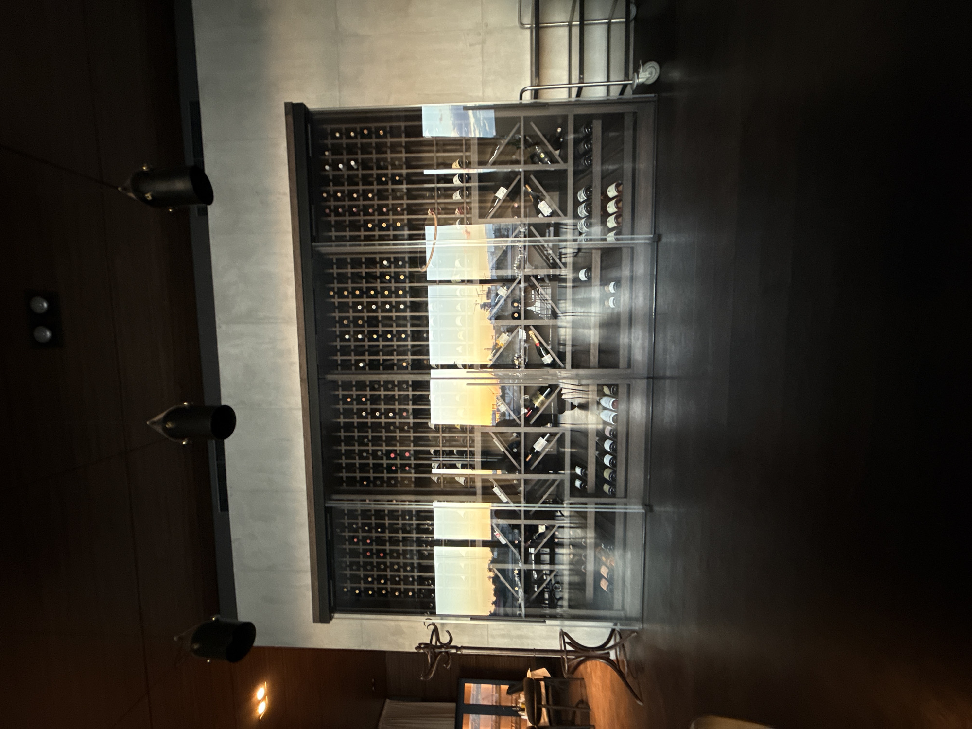 The image shows an indoor view of a wine cellar in Belgrade, Serbia. The cellar is enclosed by a metal-framed glass structure containing rows of wine bottles. A metal clothing rack with wheels is visible near the top, and a coat rack and chair are at the bottom. There are pendant lights hanging from the ceiling. A distorted reflection of a city skyline can be seen through the glass doors of the wine cellar.