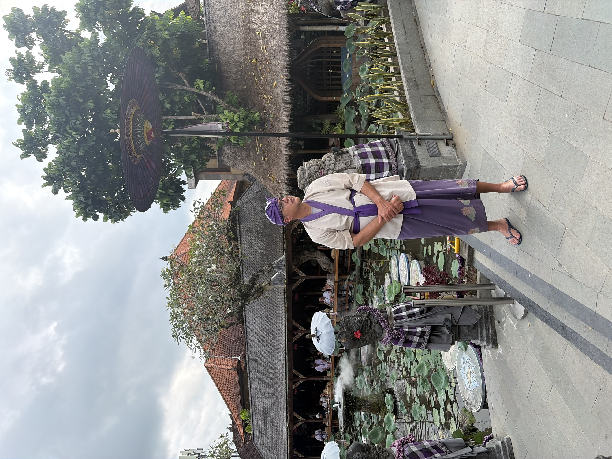 In the city of Ubud District, Indonesia, a man stands on a paved walkway beside a pond filled with water lilies. He is wearing traditional Balinese attire, consisting of a beige robe tied with a purple sash and a matching purple sarong. He has glasses and a purple head covering. In the background, there are several buildings with thatched roofs and red tile roofs, and a large tree. Two stone statues are partially visible on either side of the man. One statue is adorned with a purple and white checkered cloth and a red flower. The pond has a small fountain in the center, and several circular decorative platforms are submerged in the water. The sky is overcast.