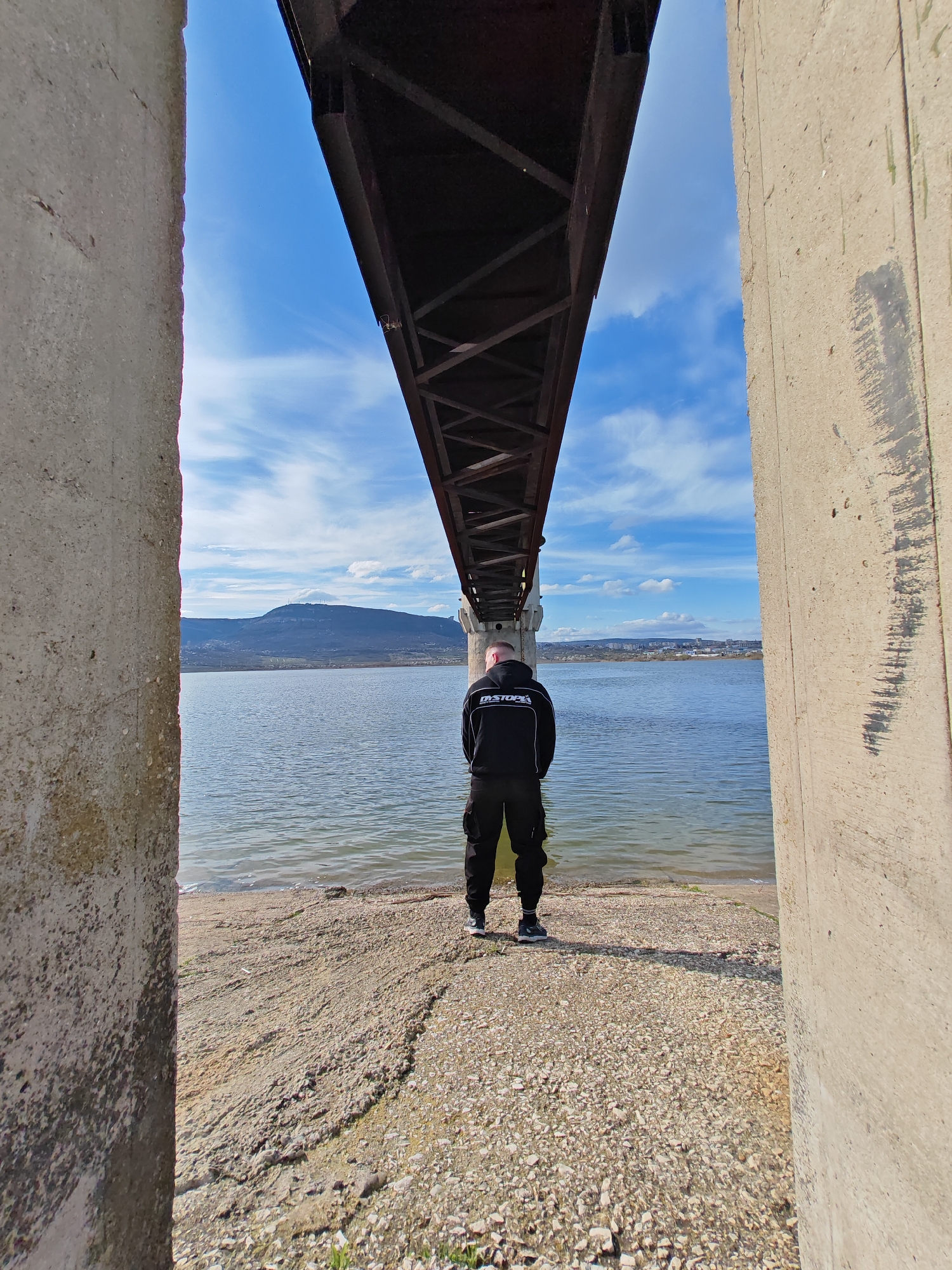 A man stands facing a body of water framed by two concrete pillars and a bridge structure overhead. The man is wearing a black tracksuit with white lettering on the back. He is standing on a rocky shore, and beyond the water is a mountain range and the city of Shumen, Bulgaria. The sky is blue with scattered clouds. The scene is under a bridge, likely a railway bridge, on the shore of Lake Shumen.