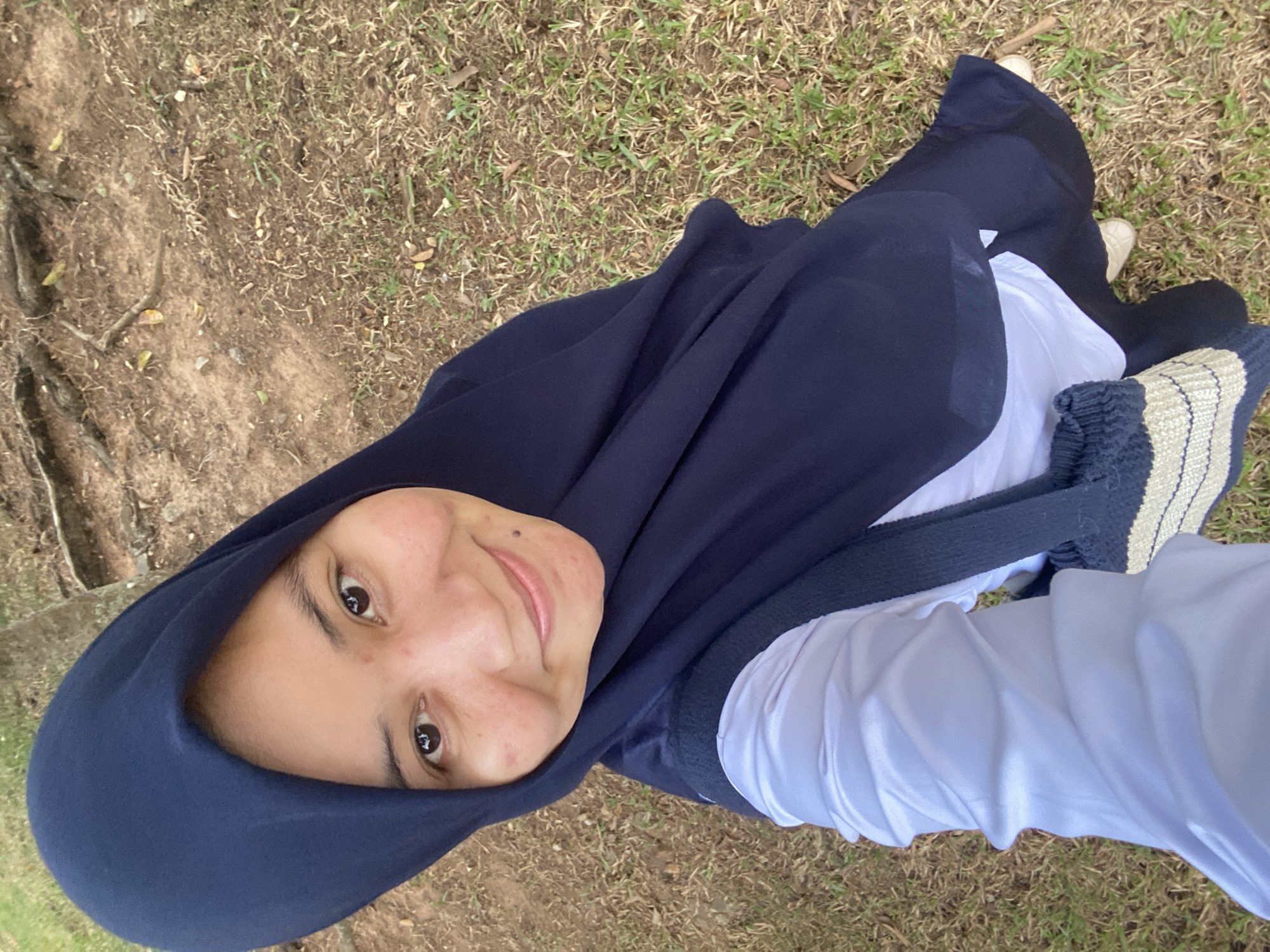 A young woman wearing a navy blue hijab and a white shirt stands outdoors, holding a striped bag. She is looking towards the camera and smiling. The background consists of dry grass and tree roots, suggesting an outdoor location. The image appears to be a selfie taken in a park or natural area within Banjarbaru, Indonesia.