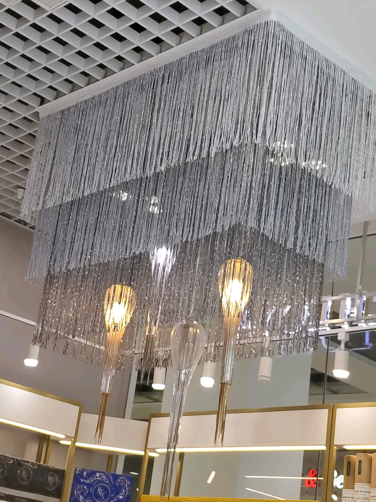 The image shows a modern chandelier with multiple tiers of silver fringe hanging from a white tiled ceiling. The chandelier has several light fixtures, some with exposed bulbs that appear to have an amber glow. The background suggests an indoor commercial space, possibly a store, with displays of products on shelves and recessed lighting. There are no people visible in the image. The location is not specified in the image.