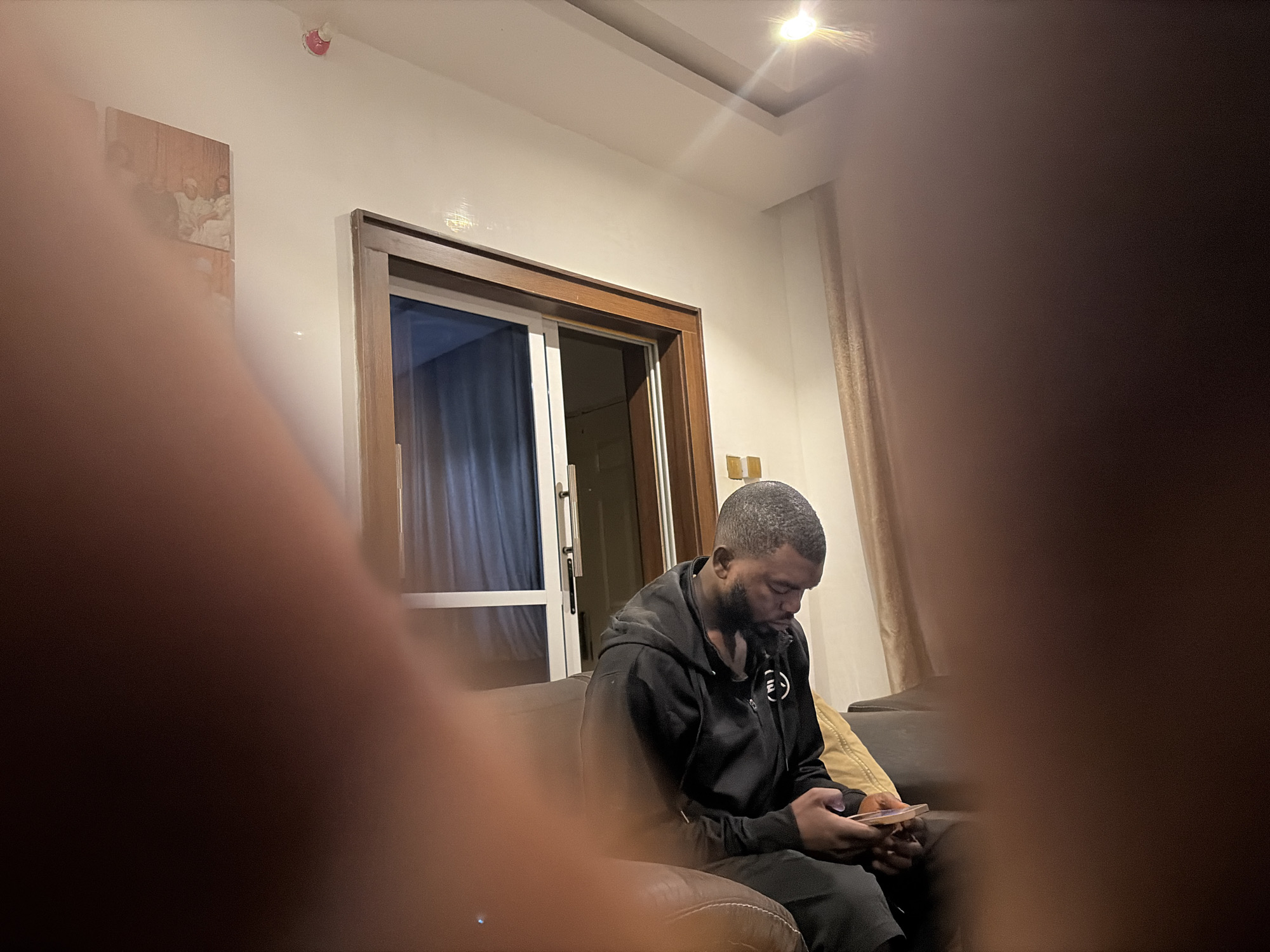 A medium indoor shot shows a person sitting on a couch. The person is an African male with a short haircut and a beard. He is wearing a black hoodie and black pants, and he is looking down at a phone he is holding in his hands. The couch is brown leather. In the background is a window with curtains. On the wall is a framed picture and a light fixture. The photograph appears to be taken in a private residence. The city is Jos, Nigeria. The act seems to be the person looking at their phone.