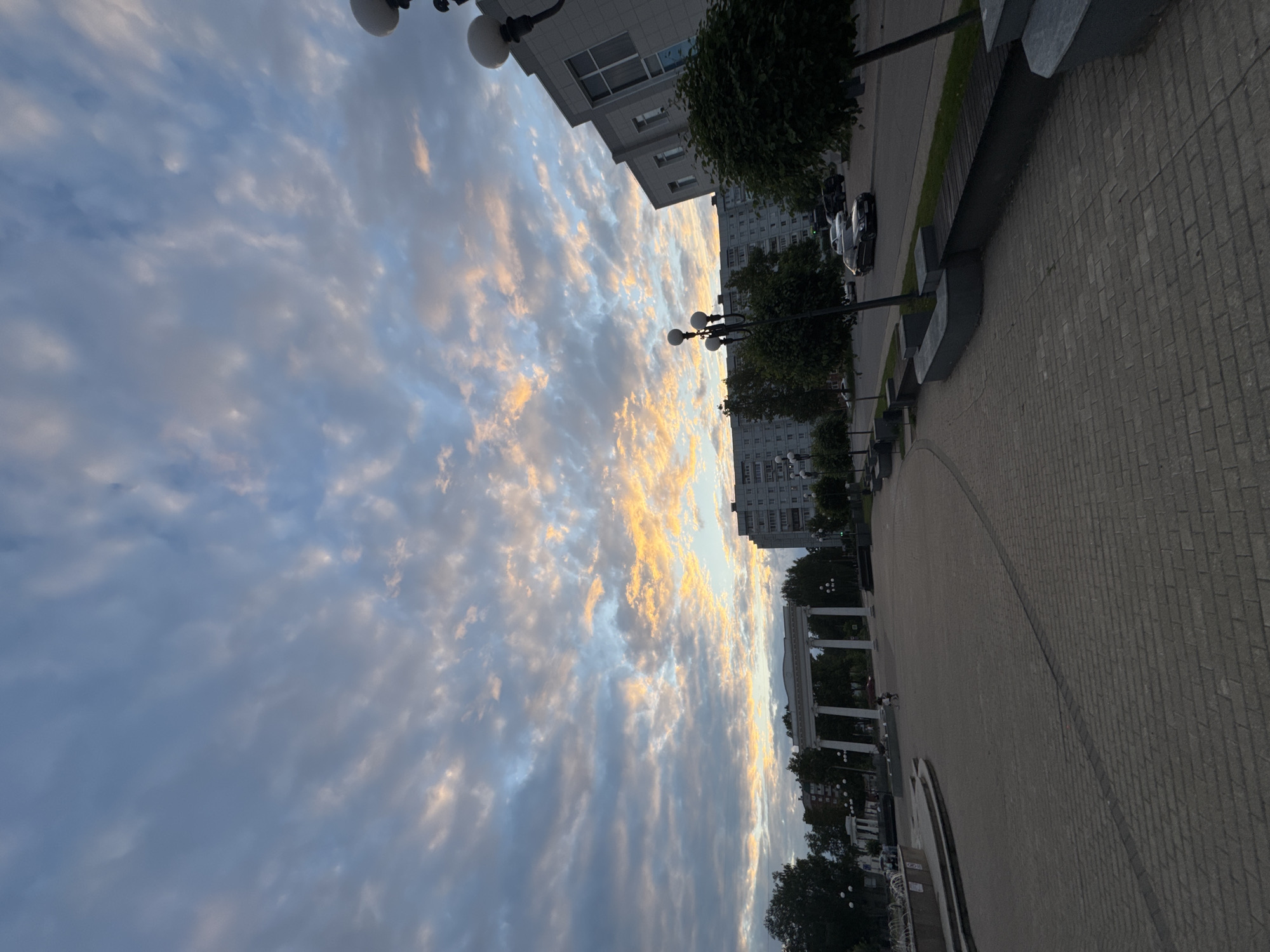 An outdoor wide-angle shot captures a city square in Chaikovsky, Russia at dusk. The sky is filled with dramatic clouds, tinged with hues of orange and yellow from the setting sun, contrasting with the soft blue and gray of the lower sky. In the background, a grand, white archway stands prominently, framed by trees. Adjacent to the archway, a multi-story residential building and a modern commercial building are visible. The foreground is dominated by a wide, paved plaza with patterned brickwork. Several lampposts with spherical lights line the plaza. On the right side of the frame, a black sedan is parked on the street next to the plaza, with other vehicles and more buildings further in the background. A few people are seen in the distance near the archway and on the left side of the plaza, near what appears to be a fountain.