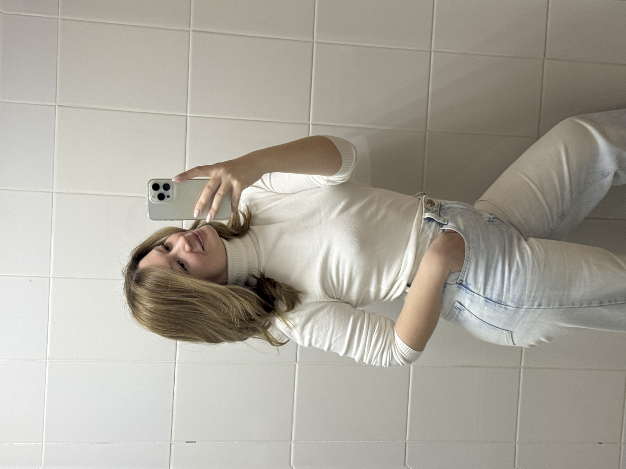 A woman is posing for a selfie in a room with white tiled walls. She is wearing a white turtleneck sweater and light-wash jeans. She is holding a phone in her left hand to take the picture. The location is likely indoors, possibly a bathroom or a similar space with tiled walls. There is no information to ascertain whether this image was captured in the city of Chaikovsky, Russia.