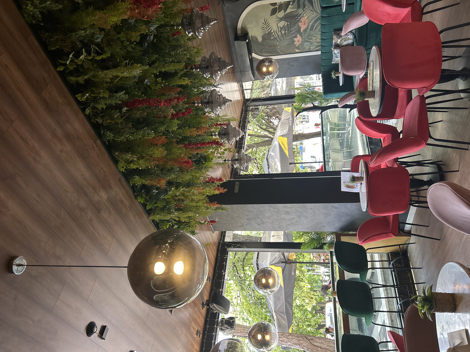 The scene depicts the interior of a modern café or restaurant located at street level in the city of Unknown, Unknown. The establishment features a distinct biophilic design with a dark wood-paneled ceiling from which extensive artificial green foliage, interspersed with red floral accents, hangs down, creating a natural canopy. Large, floor-to-ceiling glass windows line one side, offering a view of a treed outdoor area, possibly a park or street, where "Schweppes" branded umbrellas are visible, indicating an outdoor seating section. Through the windows, blurred figures of people are seen outside, some seated and others walking.
The interior is furnished with a variety of seating options. Numerous small, round tables with light-colored, possibly marble, tops are scattered throughout the space. These tables are predominantly accompanied by red velvet chairs with black metal legs. Along a far wall, a dark green velvet banquette provides additional seating, also paired with small tables. Near the large windows, a counter-height table or bar is equipped with tall, dark green velvet bar stools featuring black metal frames. Some pink velvet chairs are also visible. Many tables display small potted plants, resembling succulents, and menus or promotional items.
Overhead, multiple types of pendant lighting illuminate the space: large, spherical, smoky-grey glass pendants with visible bulbs are centrally featured, and a series of smaller, silver-colored, tiered conical pendants hang along a parallel line. Black overhead speakers are mounted on the ceiling. A white "EXIT" sign is affixed to a black wall section. The walls are a mix of dark grey textured surfaces and black structural elements. An arched wall art piece depicting a tropical jungle scene with pink flowers and green foliage is prominently displayed on one wall. An air conditioning unit is also mounted high on a wall. No specific individuals or interactions are clearly discernible within the indoor area of the primary view.