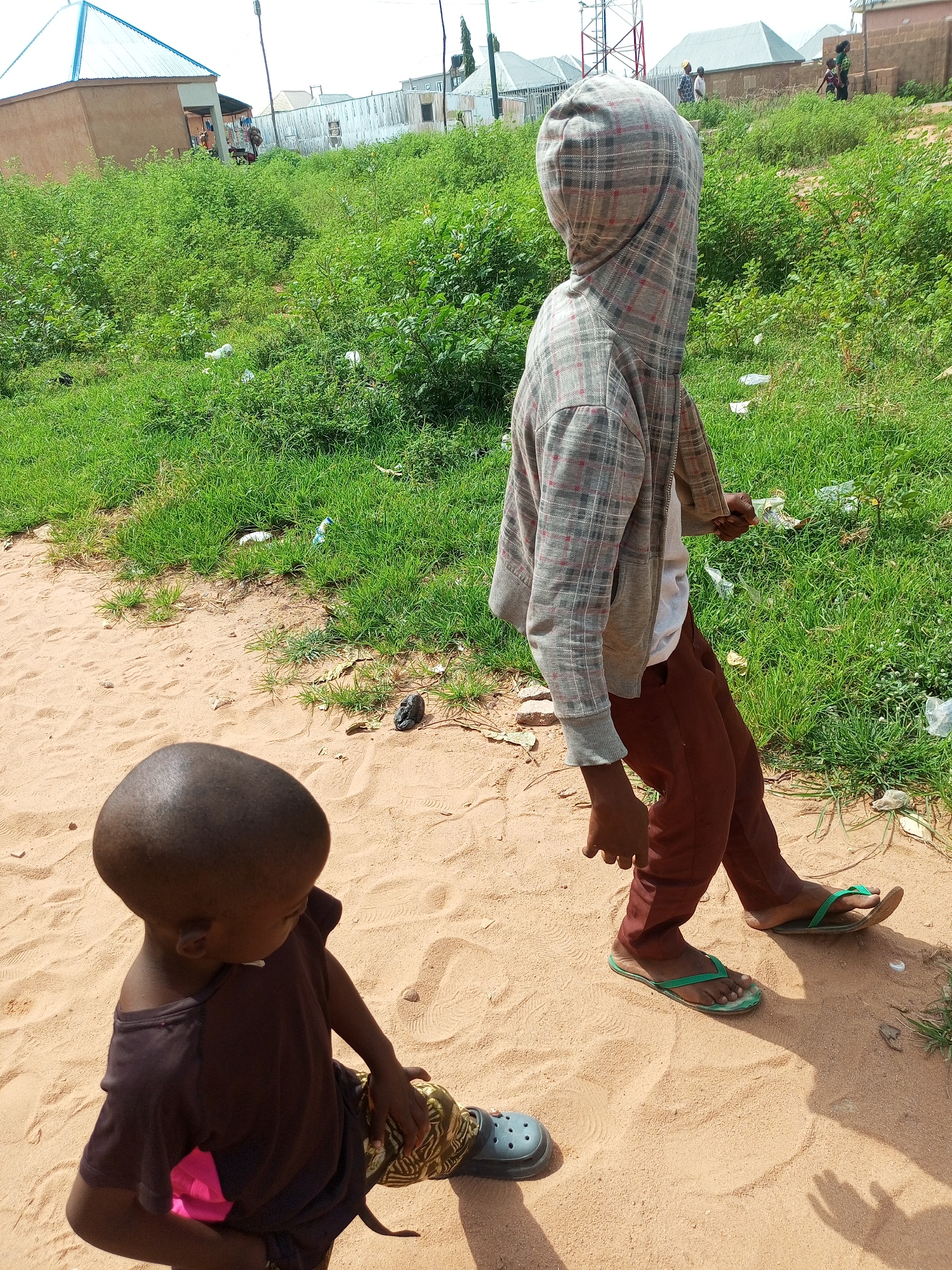 In the city of Tempure, Nigeria, on a dirt path beside a grassy area with scattered debris, two young individuals are observed. In the foreground, a child with a shaved head wears a dark brown t-shirt and patterned pants, with a bright pink object tucked into their waistband and grey shoes on their feet. They are sitting on the sandy ground, looking down. Behind them, a slightly older child, facing away from the camera, is wearing a hooded plaid jacket over a light-colored shirt and brown pants. They are walking on the dirt path, wearing green flip-flops. In the background, several buildings with corrugated metal roofs and some vegetation are visible under a bright sky.