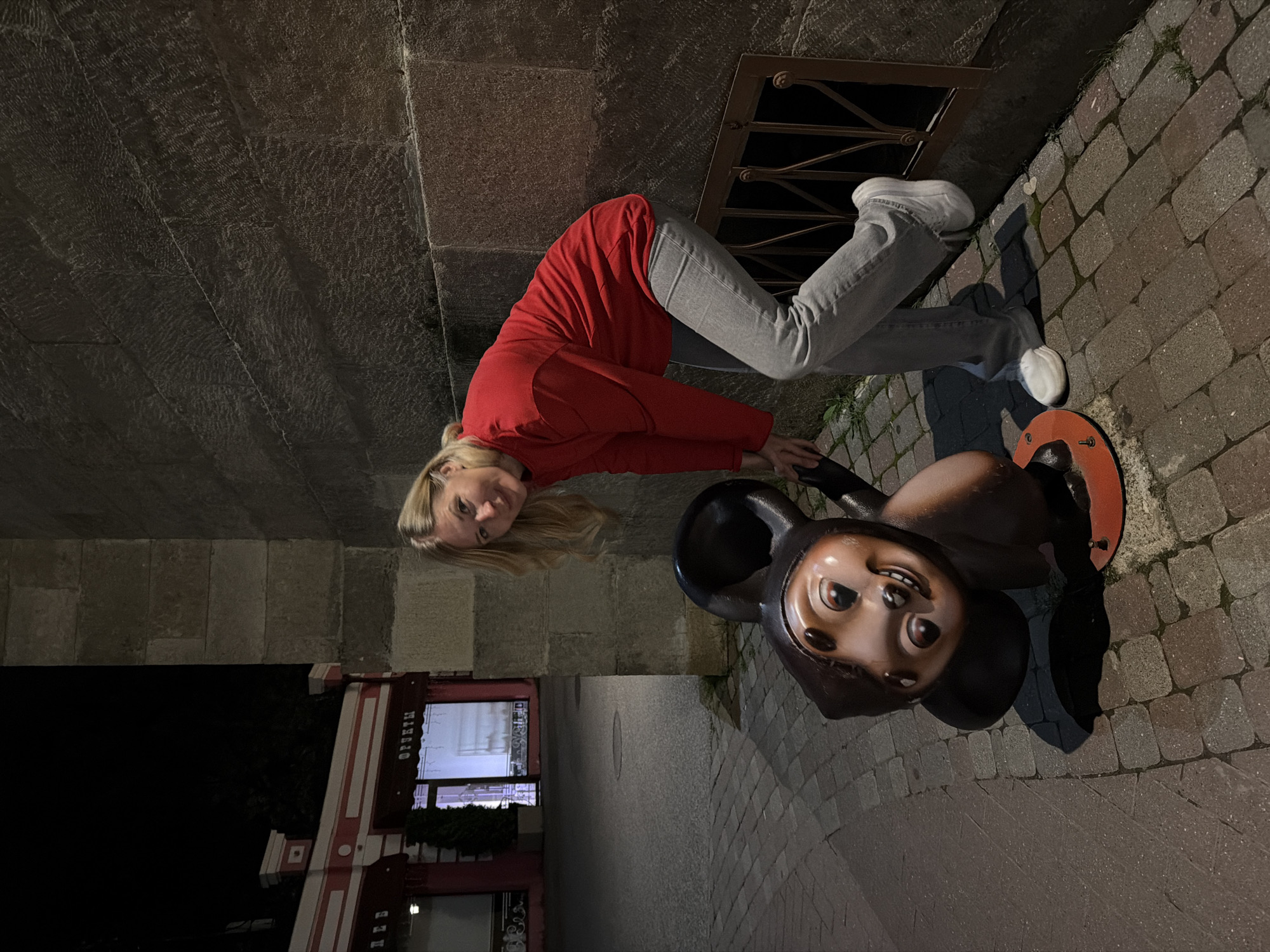 A woman with blonde hair, wearing a red long-sleeved shirt and gray jeans, is posing with a Cheburashka statue on a cobblestone street at night. The statue is a brown, cartoonish character with large ears and a wide smile. The woman is leaning against a stone wall, with her hand on the statue. In the background, there is a building with red and white trim, and shop windows. The image appears to have been taken in Kislovodsk, Russia.