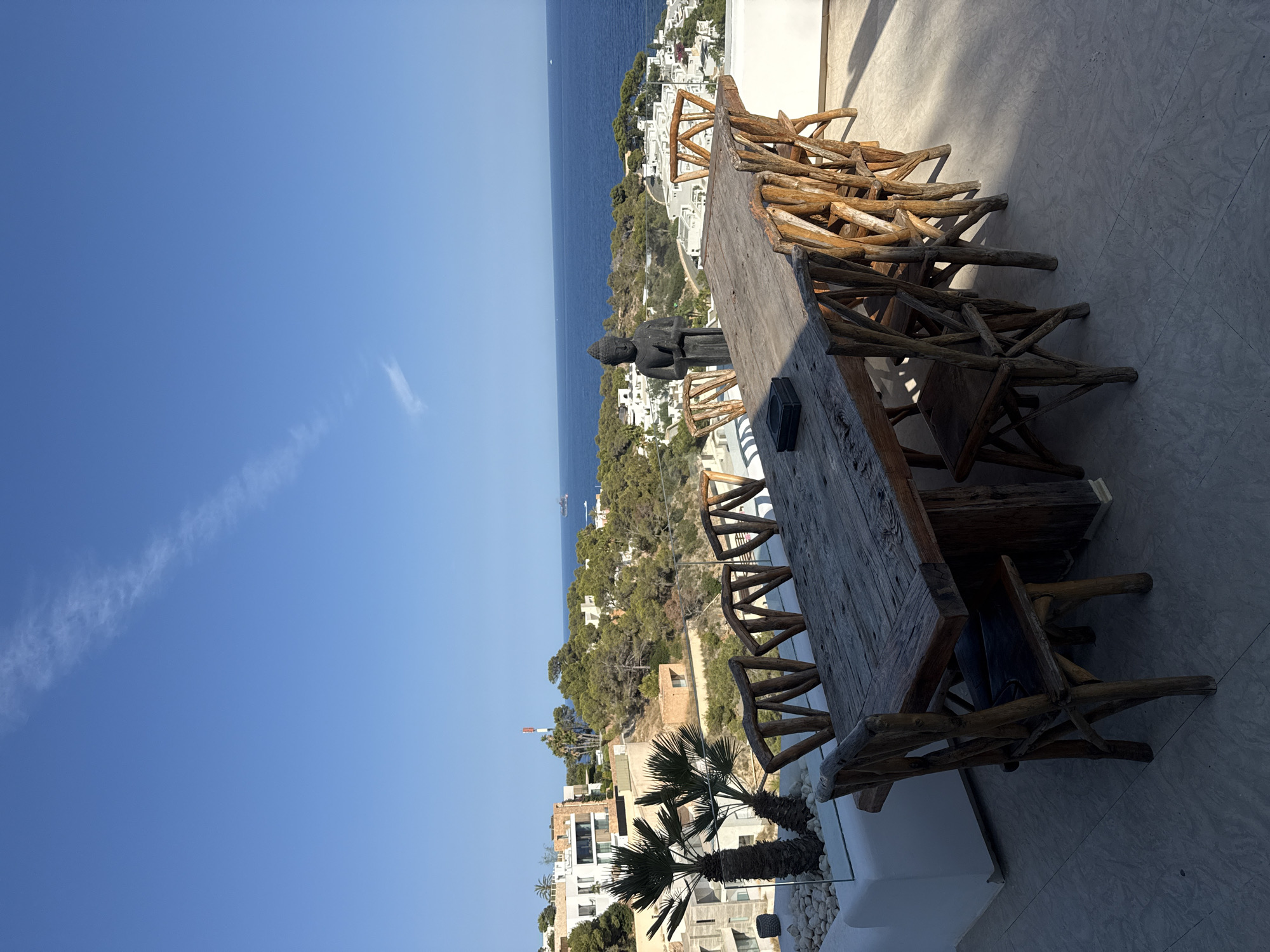 This image depicts an outdoor dining area with a rustic wooden table and chairs set on a tiled patio, overlooking a coastal landscape in Santa Eulària des Riu, Spain. The table, constructed from weathered, reclaimed wood, is large enough to seat at least eight people, with intricately carved wooden chairs arranged around it. To the left of the table, a minimalist white planter holds two palm trees. A dark, stone statue of Buddha stands behind the table, facing the sea.
In the background, a lush green hillside dotted with white buildings, characteristic of Mediterranean architecture, slopes down towards the vibrant blue Mediterranean Sea. The water is calm, with a small boat visible in the distance. The sky is a clear, bright blue with a faint, wispy cloud streak. A red and white telecommunications tower is visible on the hilltop. The scene is bathed in bright sunlight, casting long shadows from the table and chairs onto the patio. The overall impression is one of a serene and luxurious outdoor living space with a stunning natural vista.