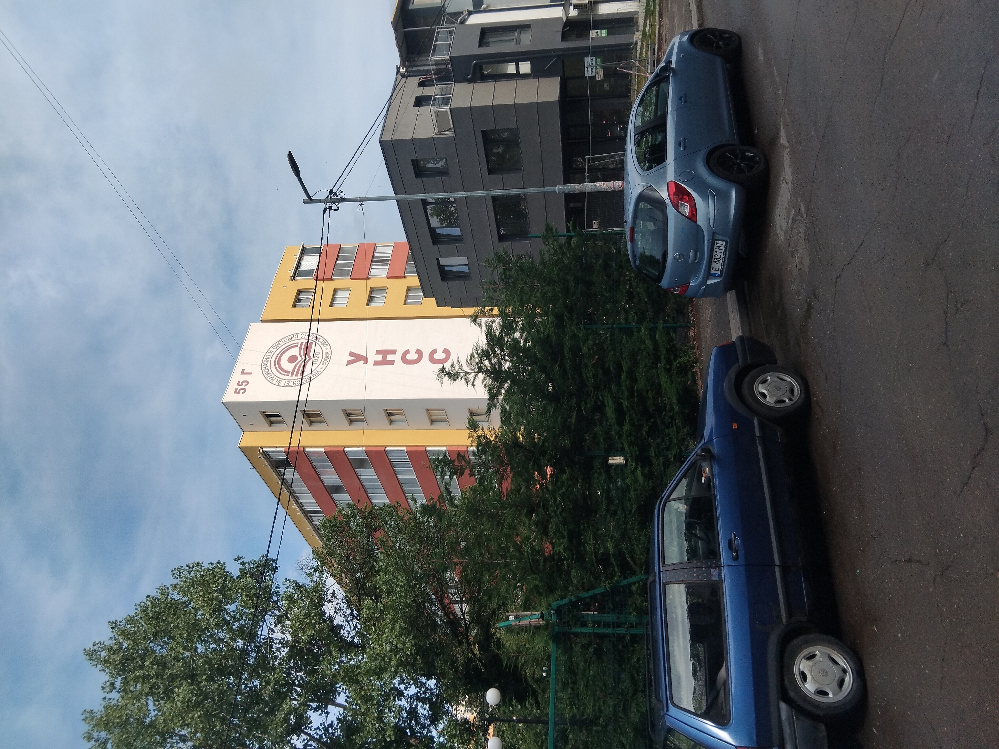 The image captures an urban street scene in Sofia, Bulgaria, featuring buildings, vehicles, and greenery under an overcast sky.
**Subjects:** In the foreground, two cars are parked on a paved street. The prominent car on the right is a blue hatchback (likely an Opel Corsa D) with the Bulgarian license plate "E 4831 HT." To its left, partially obscured by trees, is the front portion of a darker blue or dark gray older model hatchback. Several trees and bushes are visible, including dense green foliage that partially hides the buildings.
**Buildings:** Two distinct buildings dominate the background. On the left is a tall, multi-story building with a facade featuring horizontal stripes of red and yellow, topped by a large white section. To its right, a modern-looking, multi-story building with a dark gray or black facade and rectangular windows stands behind the blue car. A utility pole with wires stretches across the upper part of the image.
**Activity or Event:** The scene depicts a quiet urban environment with parked cars, suggesting a normal daytime setting with no specific activity or event taking place.
**Notable Details:** The time of day appears to be daytime, likely an overcast day, given the diffused light and lack of strong shadows. The sky is a light blue with visible clouds.
**Visible Text:** The most prominent text is on the large white section of the colorful building. Large red Cyrillic letters spell "УНСС" (UNSS), which stands for the University of National and World Economy. Above this, "55 Г" (55 G, likely "55 години" meaning "55 years") is visible, possibly commemorating an anniversary. Adjacent to "УНСС" is a circular emblem with smaller, partially legible Cyrillic text, including "УНИВЕРСИТЕТ ЗА НАЦИОНАЛНО И СВЕТОВНО СТОПАНСТВО" (University of National and World Economy) and "СОФИЯ * 1920" (Sofia * 1920), indicating the university's location and founding year. The license plate of the blue car reads "E 4831 HT".